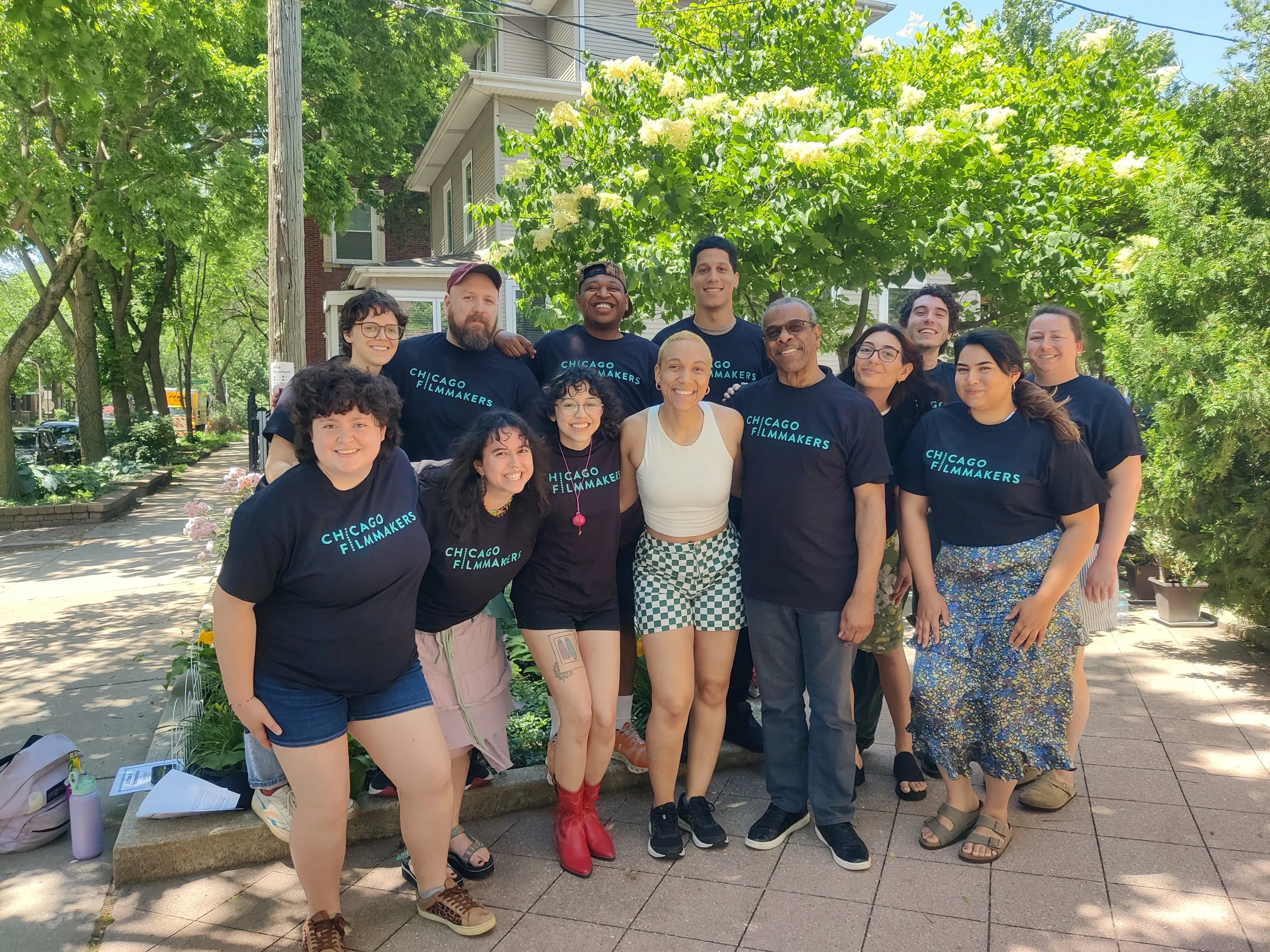 Group of diverse people smiling and posing for a photo outdoors, many wearing black T-shirts with 'CHICAGO FILMMAKERS' written on them, in a lush, green, sidewalk setting with trees and houses in the background.