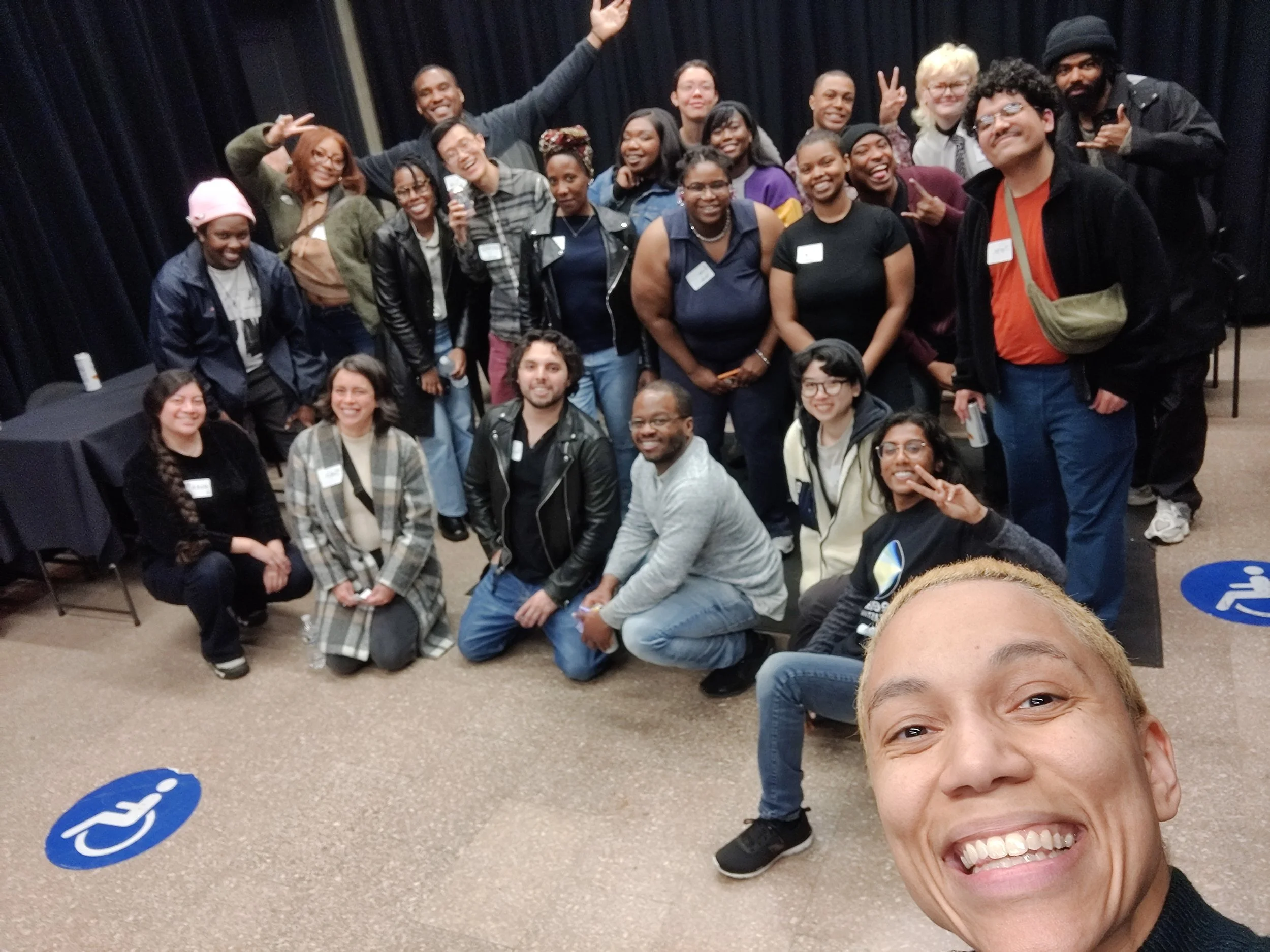 Group of diverse smiling people posing for a photo, some making peace signs, gathered indoors against a dark curtain backdrop. One person in the foreground is taking a selfie.