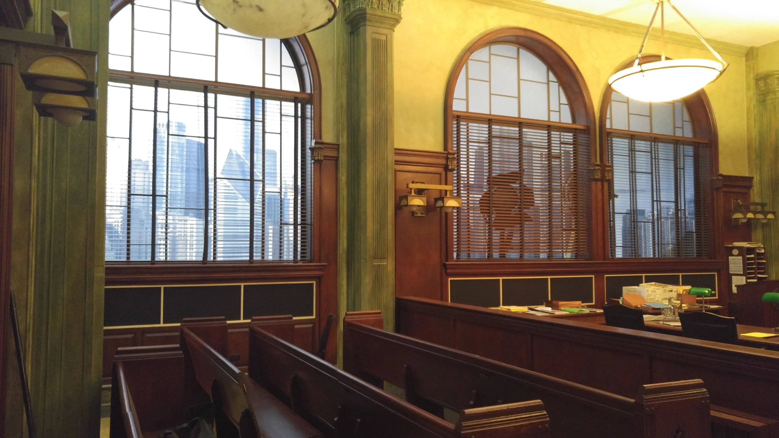 Interior of a historic courtroom or municipal building with large arched windows, wooden paneling, and overhanging lights. There are rows of wooden benches and a desk with scattered papers and office supplies.