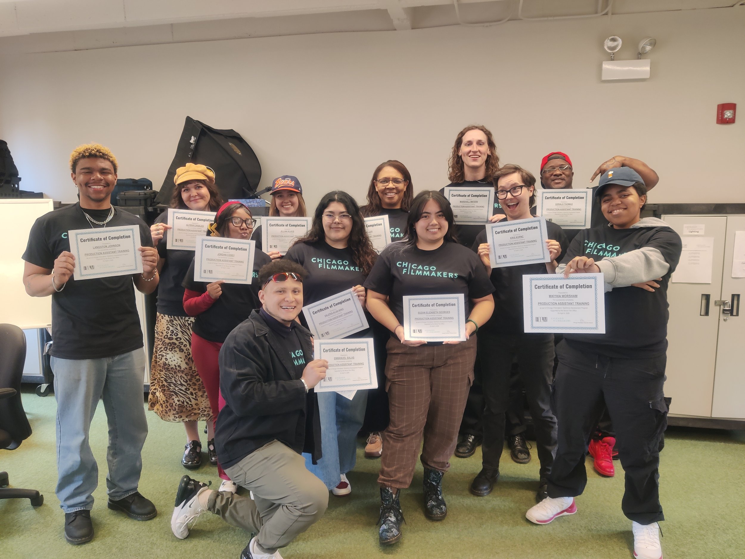 A diverse group of young adults and one older woman holding certificates of completion, smiling for a group photo in an indoor classroom or seminar room with a whiteboard and equipment in the background.