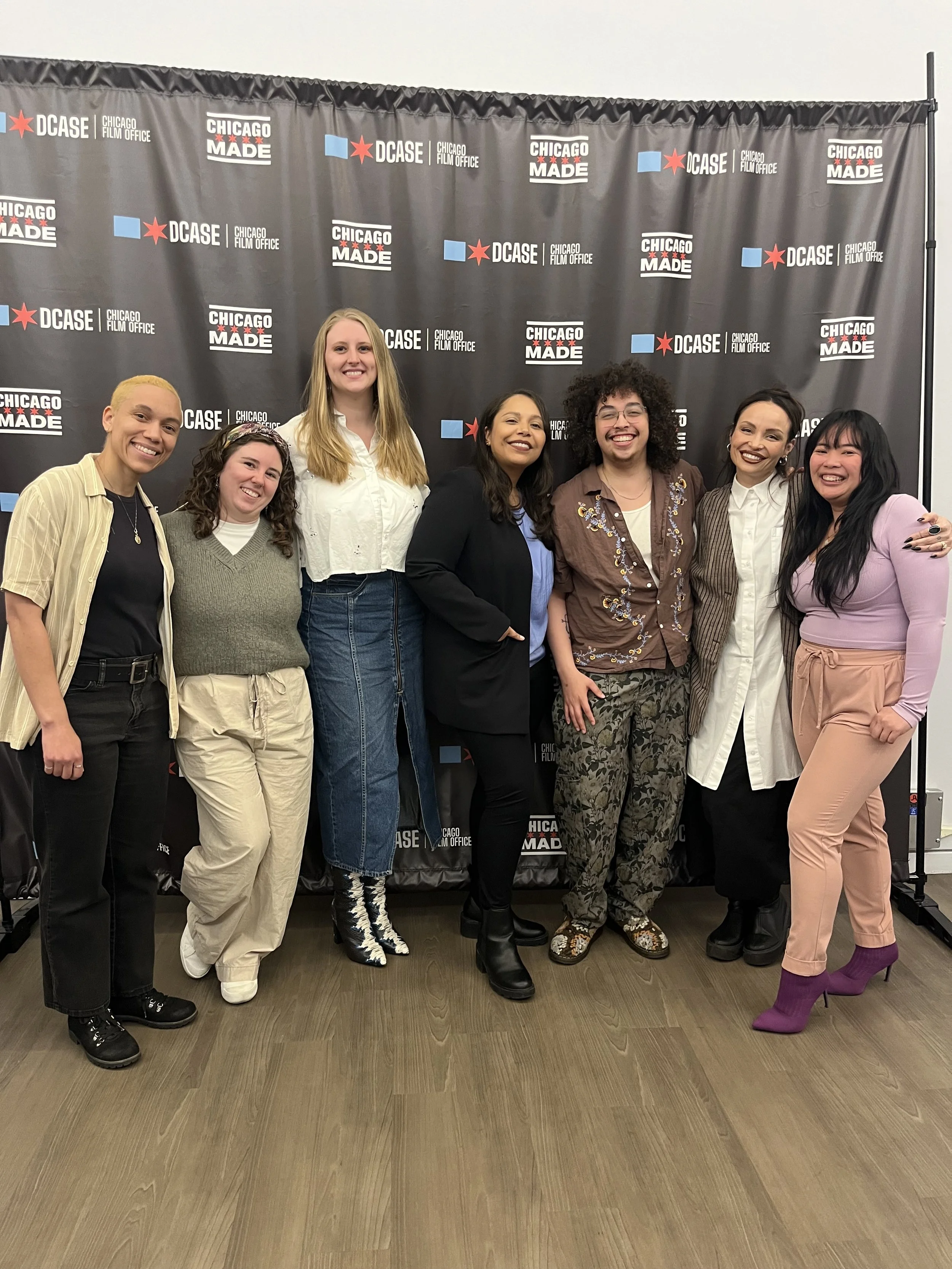 A group of seven diverse women standing together, smiling for a photo in front of a black backdrop with the words 'Chicago Made' and 'DCASE Chicago Film Office' printed repeatedly.