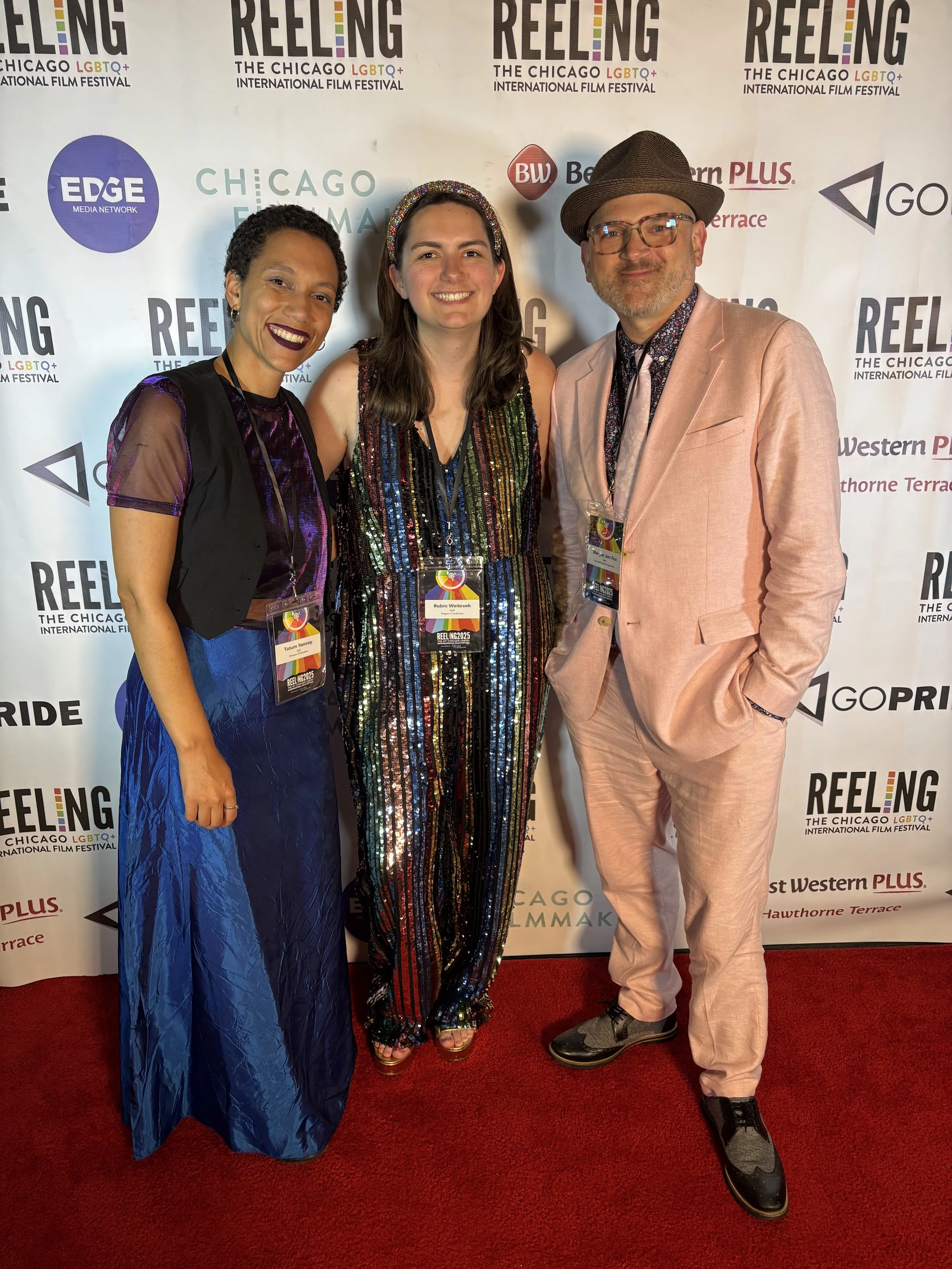 Three people standing together at the Chicago LGBTQ+ International Film Festival, smiling for the camera. The woman on the left has short curly hair, dark lipstick, and is wearing a sheer purple and black top with a long, shiny blue skirt. The woman in the middle has long brown hair, a rainbow-colored sequined dress, and a matching headband. The man on the right has glasses, a beard, a brown hat, a pink blazer, patterned shirt, and light-colored pants. They are standing in front of a step-and-repeat banner with logos and festival branding.