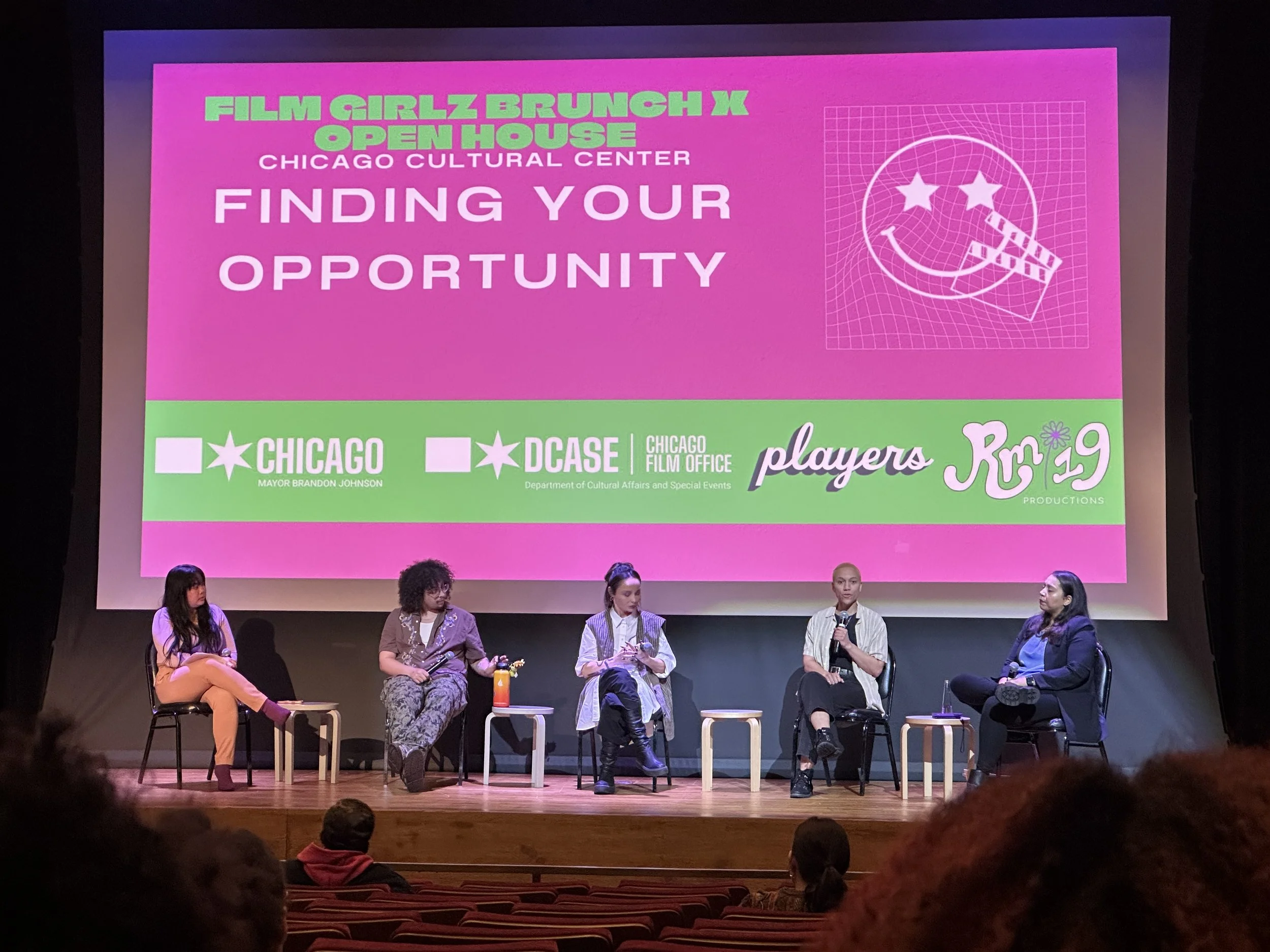 Panel discussion on stage at Chicago Cultural Center with five women seated and a large pink screen behind them displaying event details.