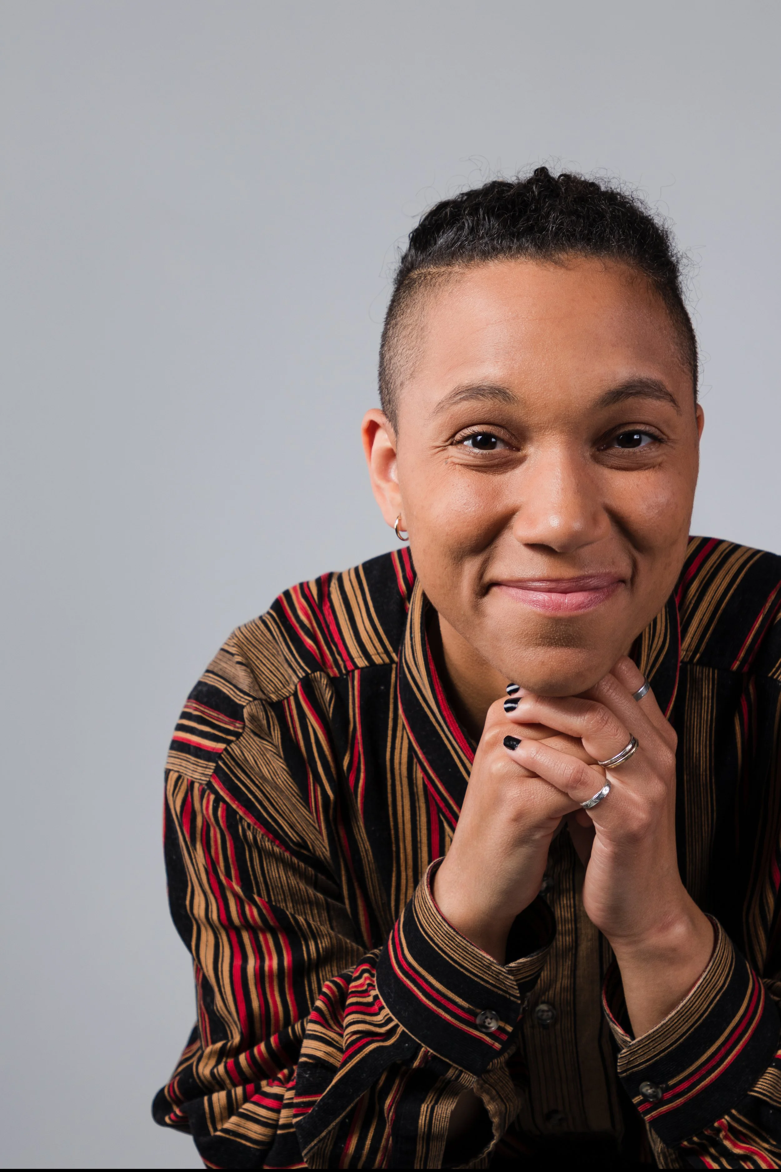 Close-up portrait of a smiling woman with short curly hair, wearing a multicolored striped shirt and rings, against a plain light gray background.
