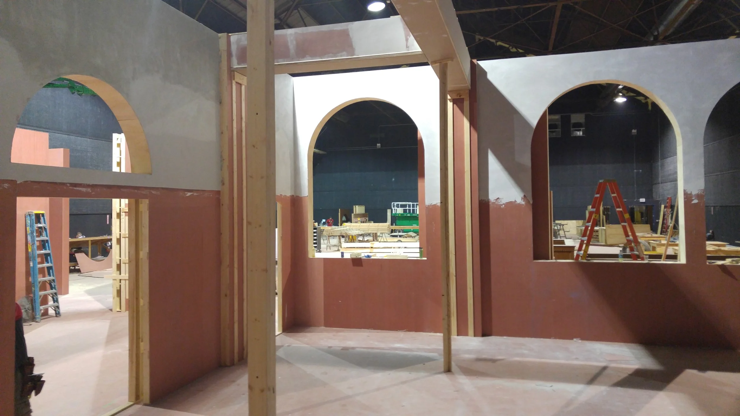 Interior of a building under construction with wooden walls and arched openings, scaffolding, ladders, and construction tools.