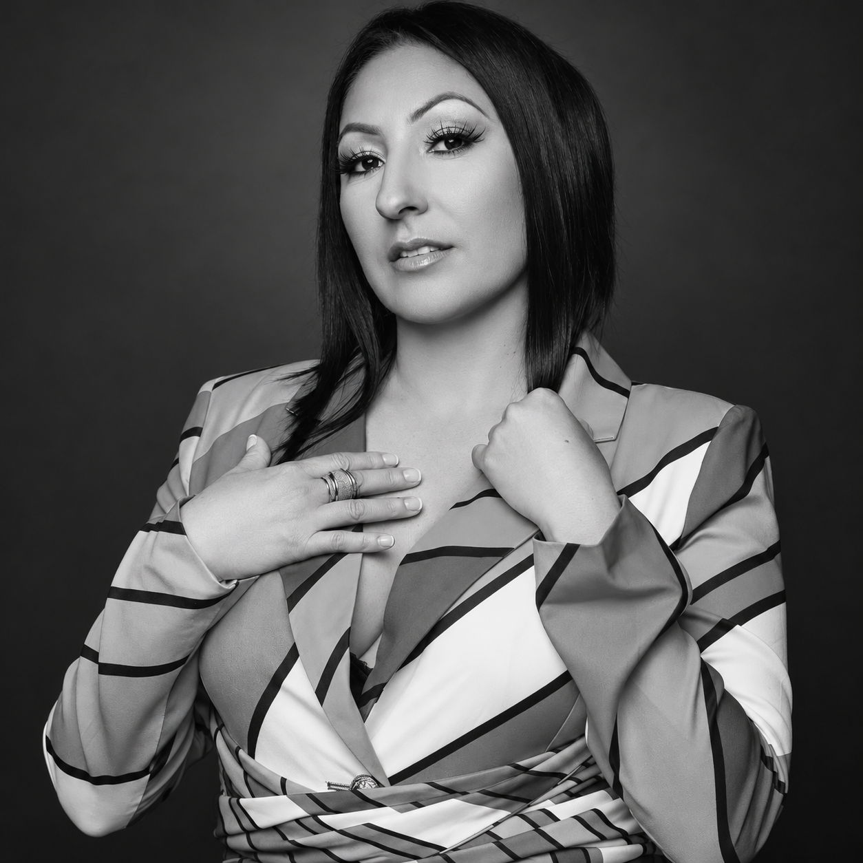 Black and white portrait of a woman with straight dark hair, wearing a striped blazer, with one hand resting on her chest and the other touching her neck, against a plain background.