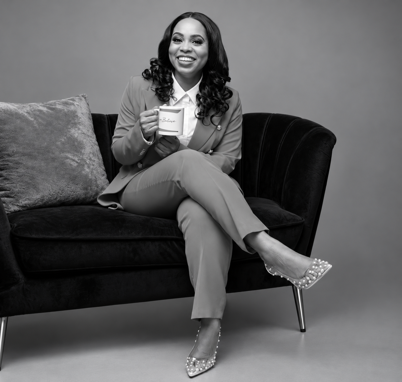 Black and white photo of a smiling woman sitting on a dark velvet sofa, holding a mug that says 'Believe', wearing a blazer, pants, and pearl-embellished high heels.