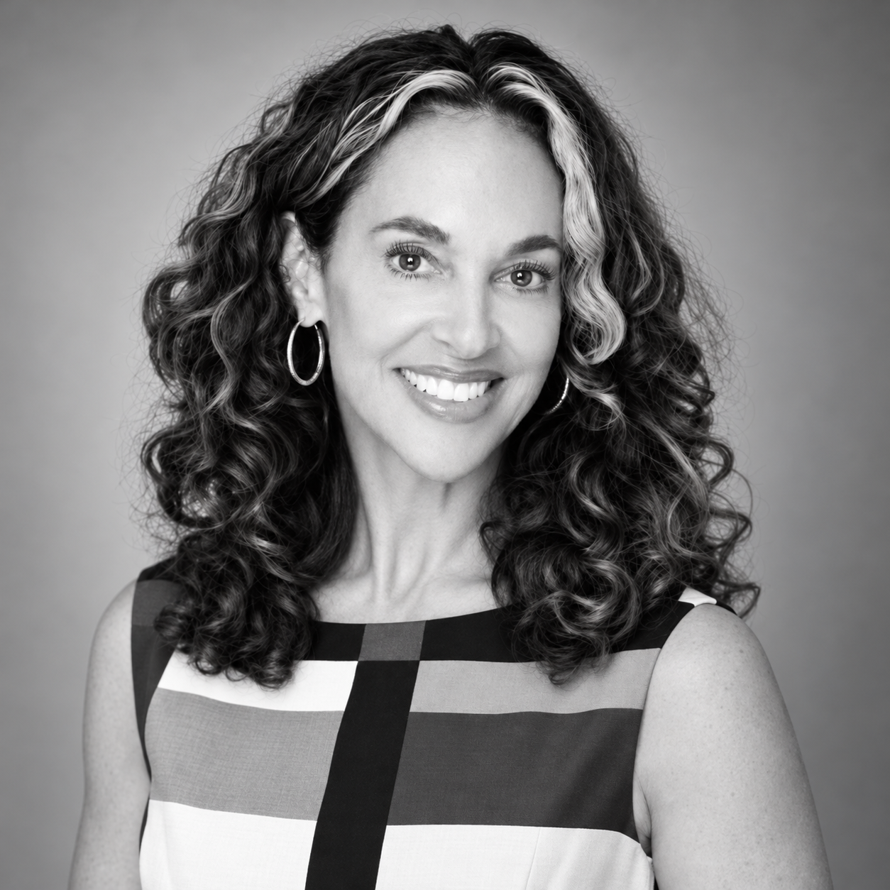 Black and white portrait of a smiling woman with curly hair, wearing hoop earrings and a sleeveless checkered top.