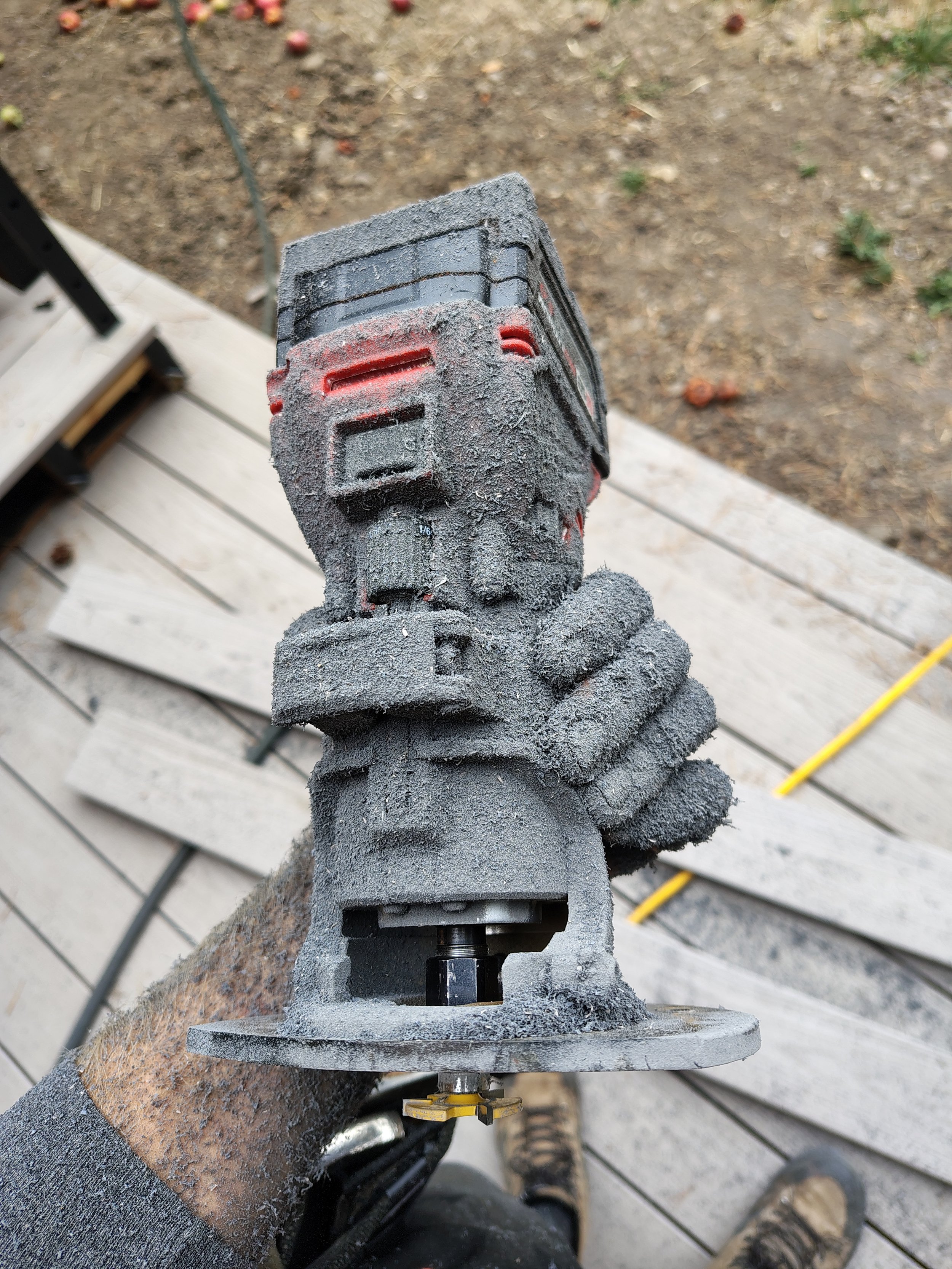 A worker's hand covered in black dust holding a power tool, possibly a grinder, with a black and red casing. The background shows a wooden deck and an outdoor area with mulch and plants.