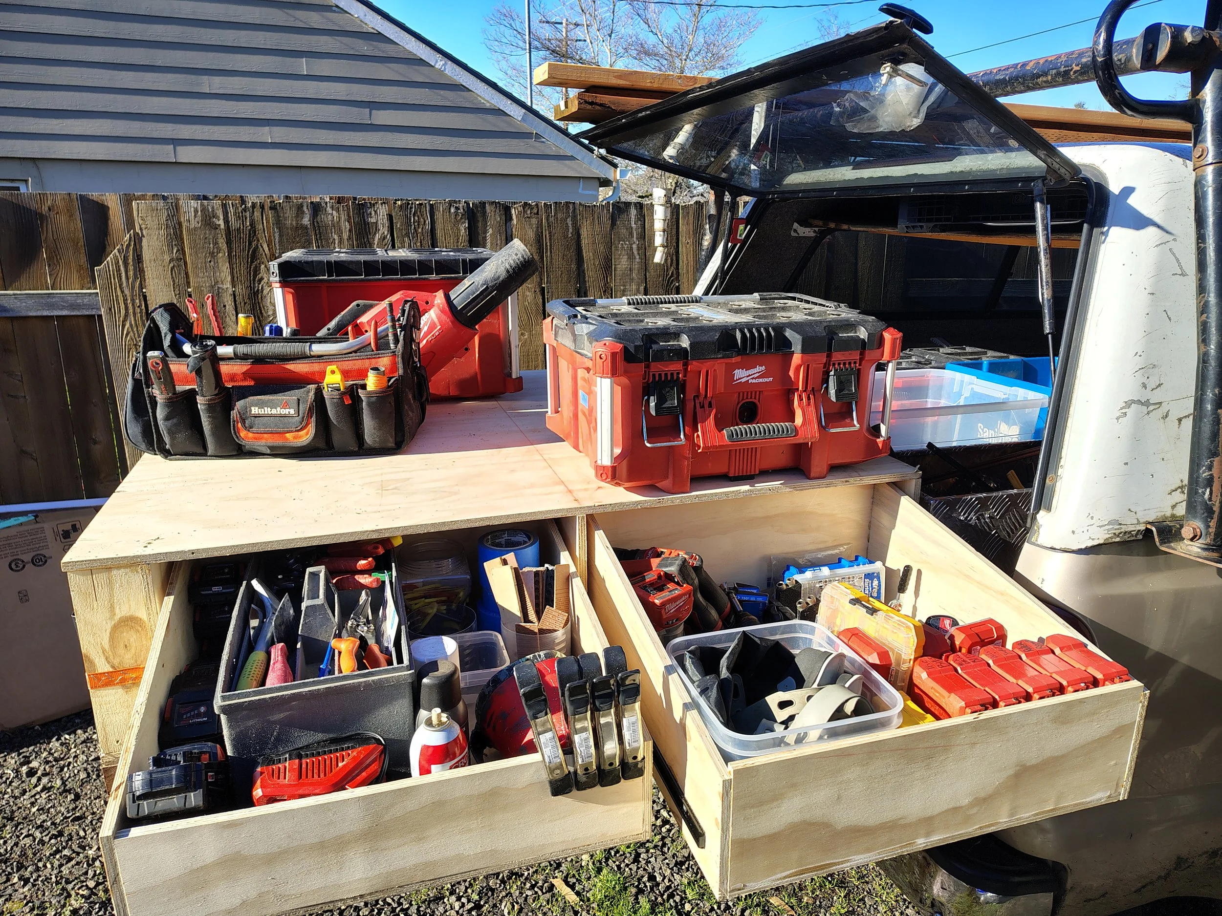 Construction project with tools and equipment on a wooden workbench outdoors, including toolbox, power drill, hand tools, and storage containers.