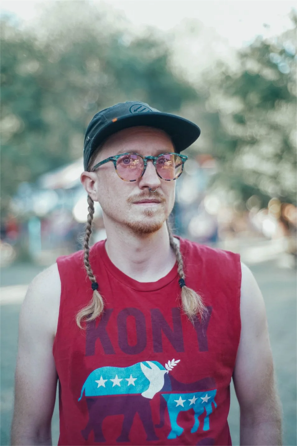 A person with glasses, braids, and a nose ring, wearing a red sleeveless shirt and a black cap outdoors.