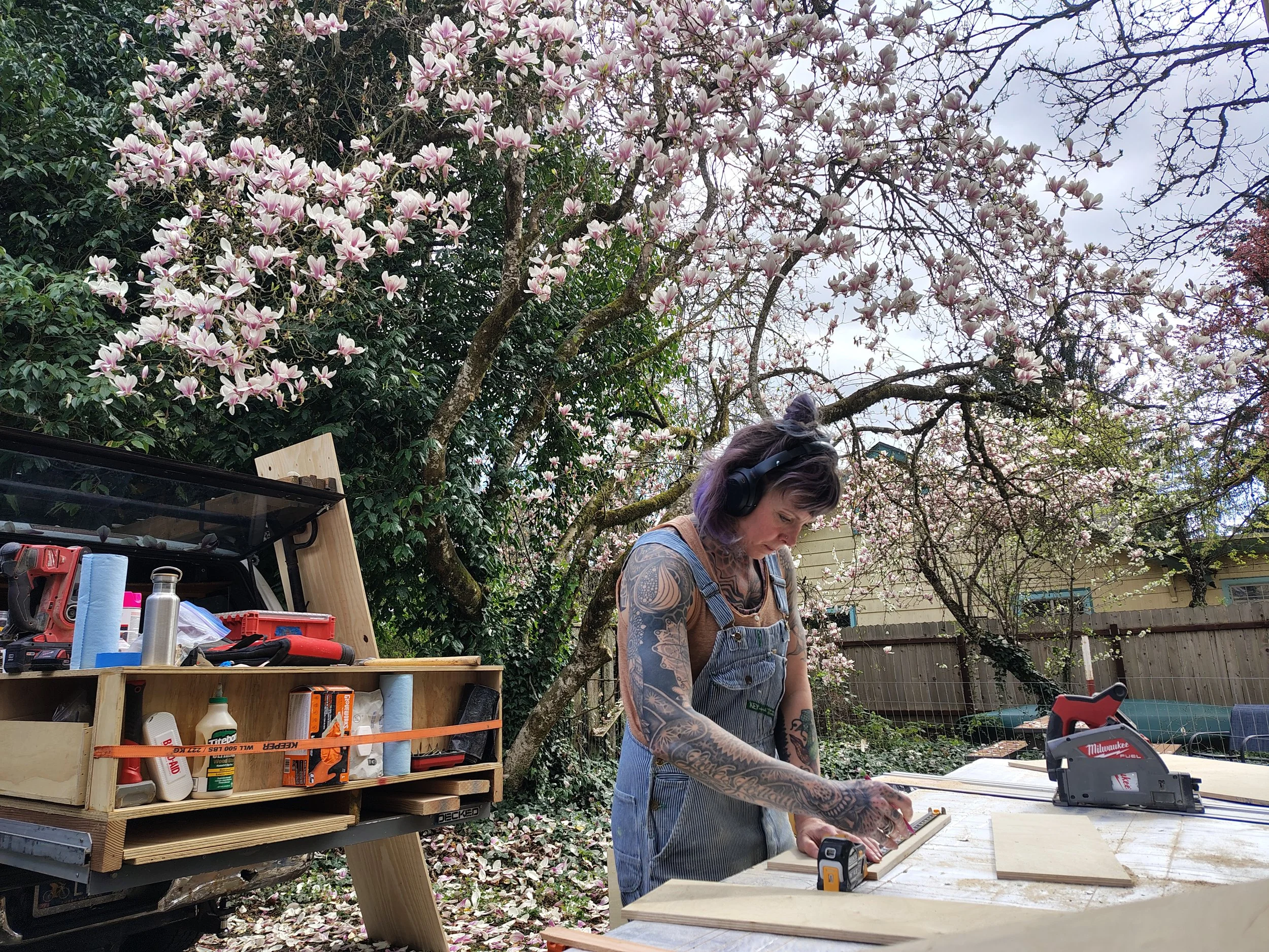 A woman with tattoos on her arms, wearing a blue striped apron, working outdoors at a wooden workbench with tools and materials, surrounded by flowering trees in a backyard.