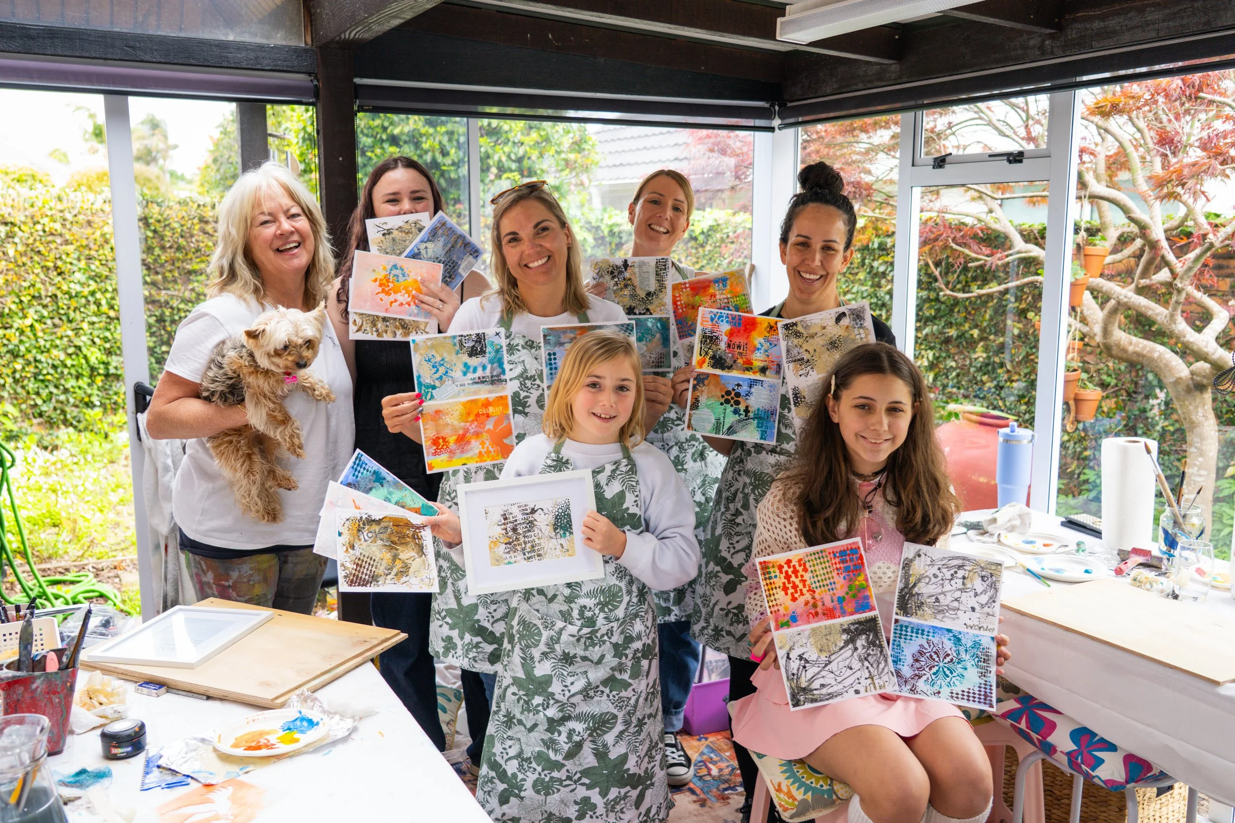 A group of women and girls holding colorful abstract art pieces at an art workshop in a sunlit indoor space with large windows and greenery outside. One woman is holding a small dog.
