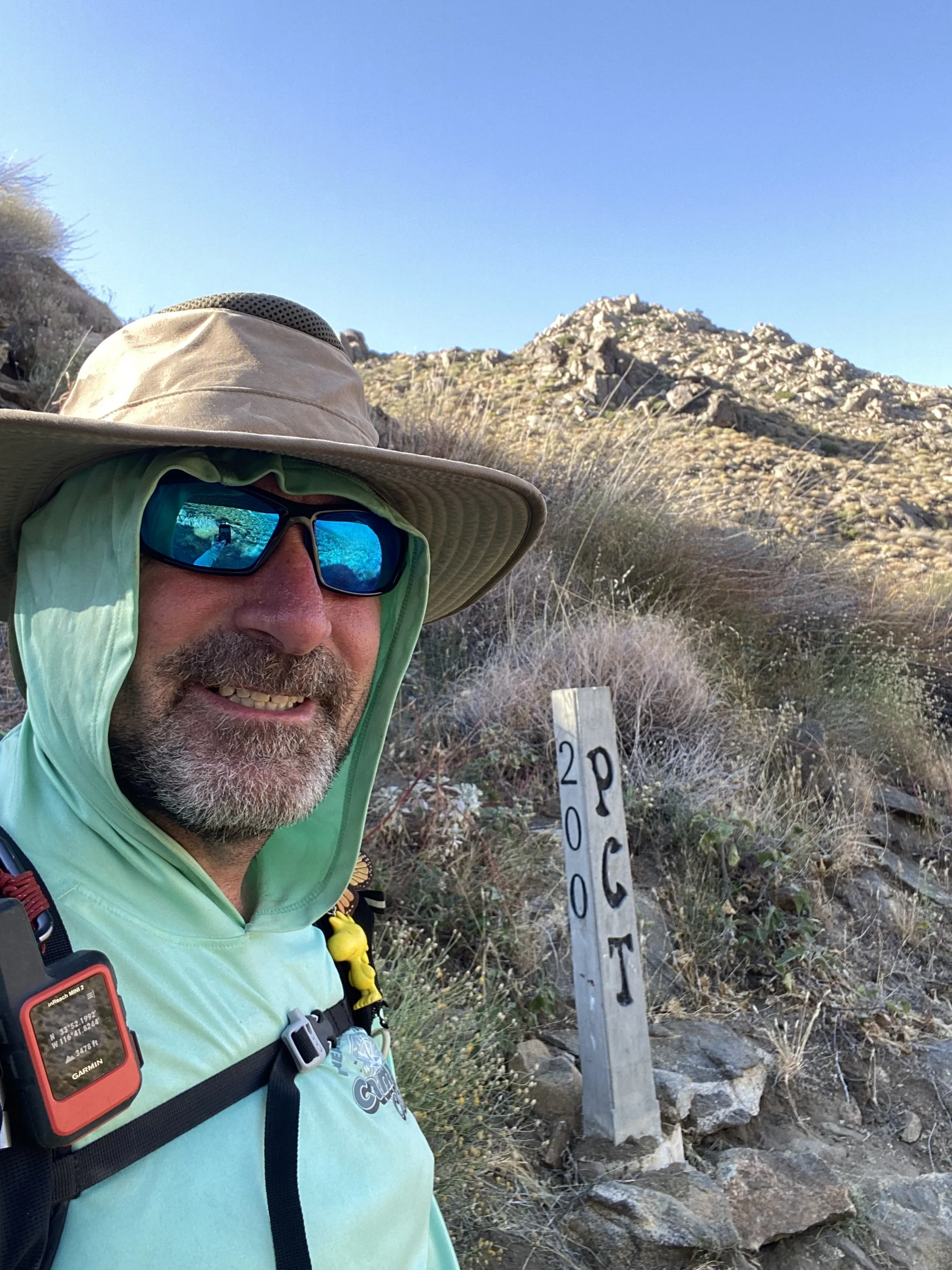 A man wearing a wide-brimmed hat, sunglasses, and a light green jacket is taking a selfie in a mountainous outdoor area. There is a trail marker with the number 209 next to him, and rocky terrain with sparse bushes behind him.