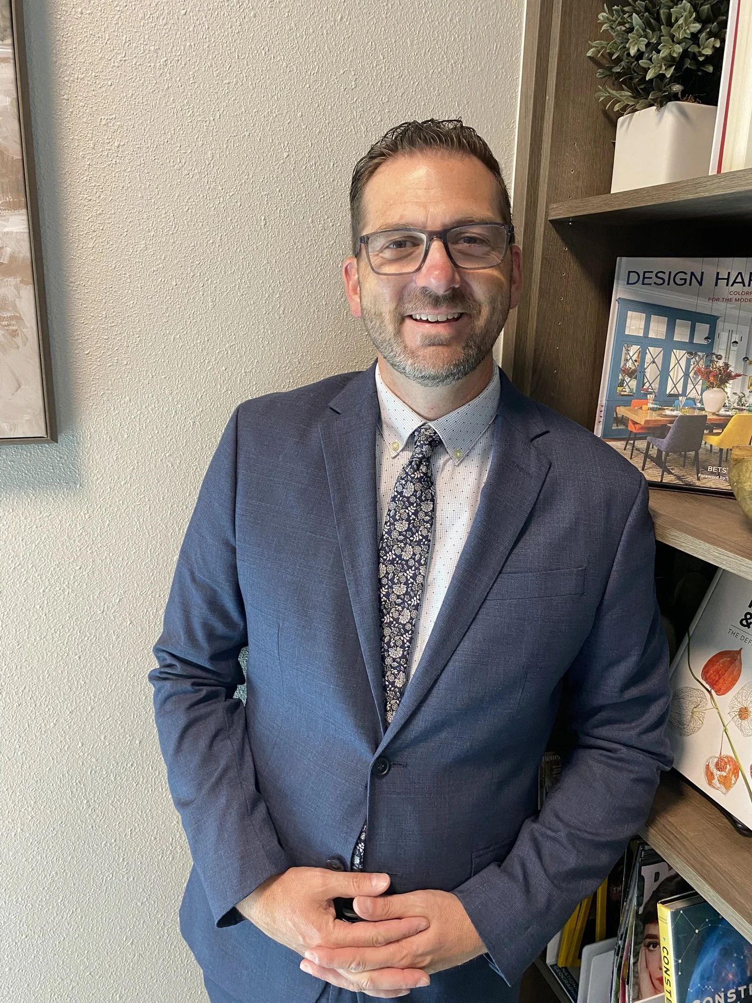 Seth Oaks in a dark blue suit, white shirt, and patterned tie standing next to a bookshelf with magazines and books, smiling at the camera.