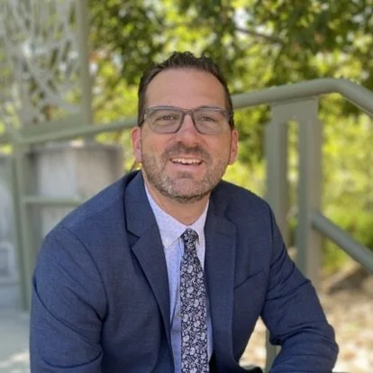 Seth Oaks wearing a blue suit, and a patterned tie, smiling outdoors with green trees and metal railings in the background.