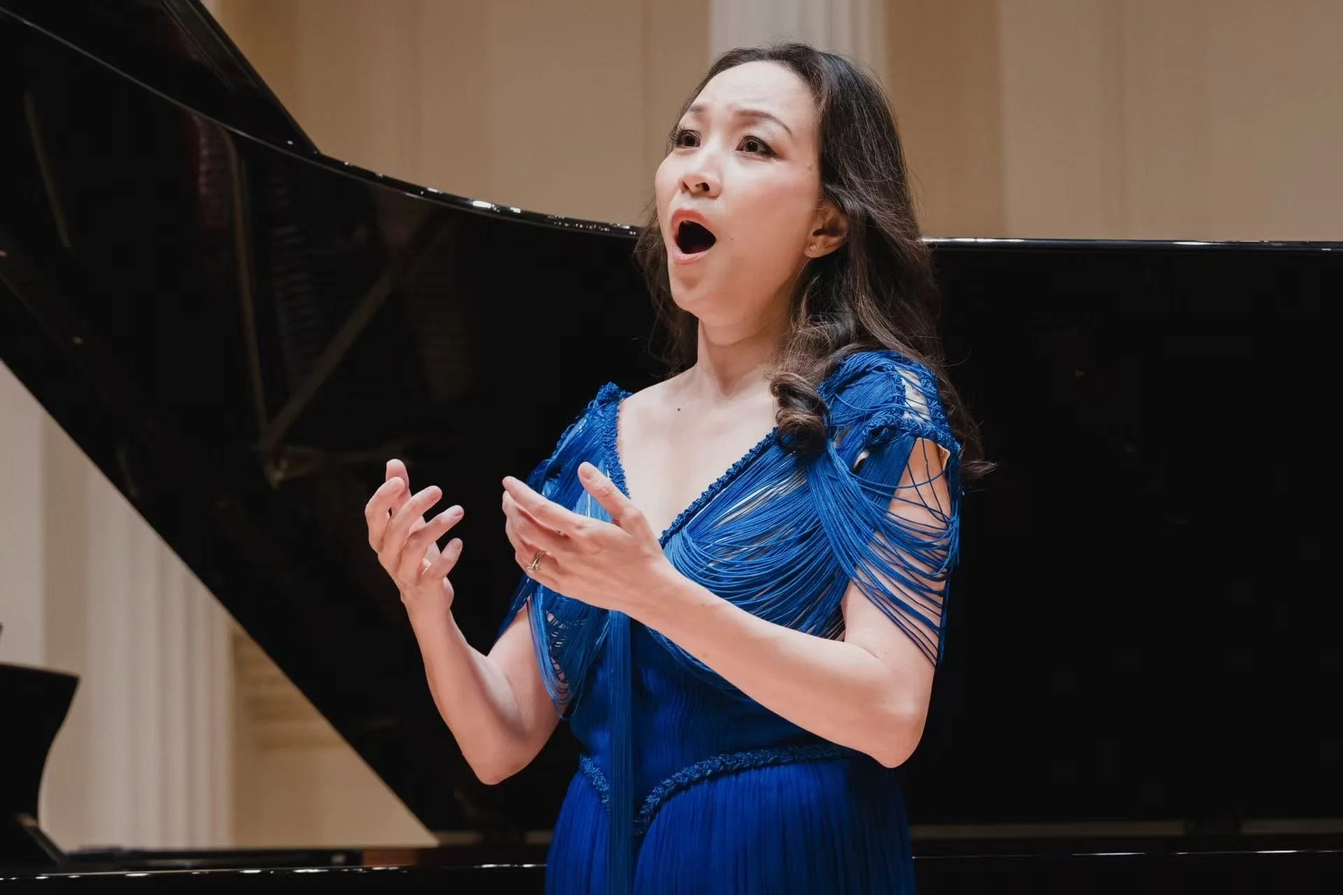 A woman in a blue dress performing passionately at a piano recital, with her mouth open as she sings or expresses emotion.