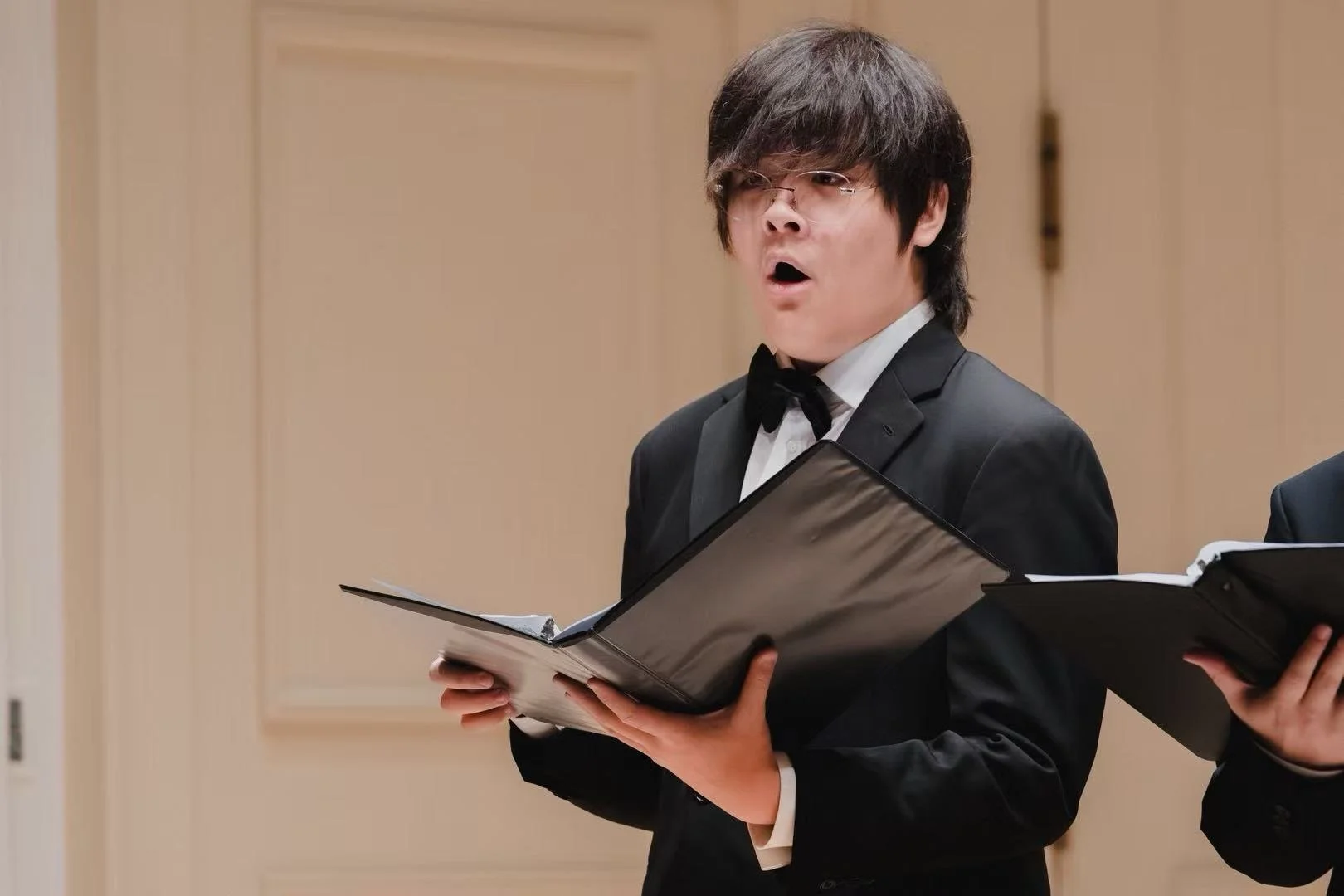 Young man wearing a tuxedo and glasses singing from sheet music in a performance