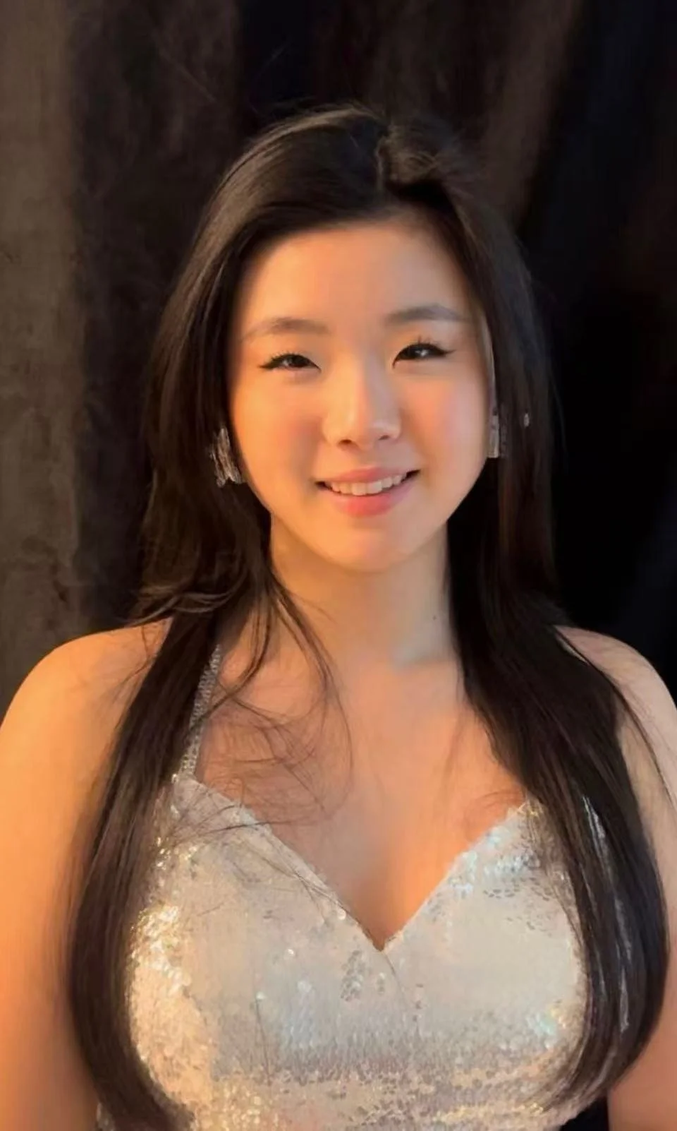 A young woman with long dark hair, wearing a silver, sequined dress and earrings, smiling at the camera.