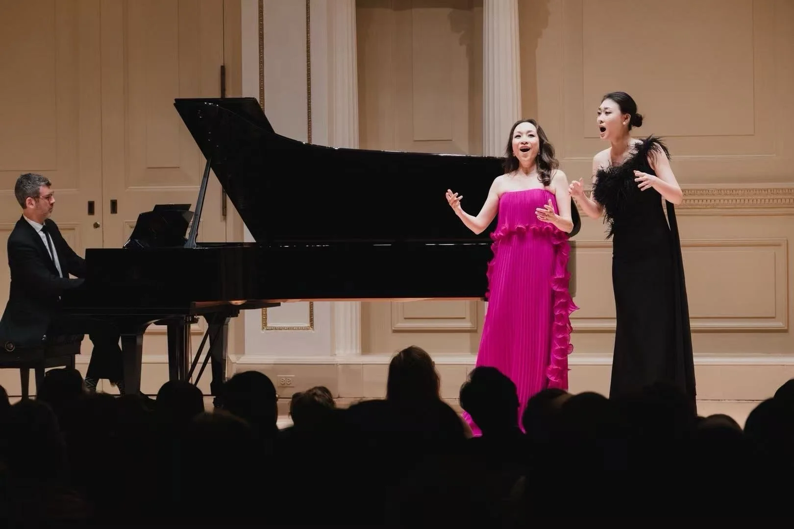 Two women singing on stage with a pianist playing a grand piano in the background, audience visible in the foreground.