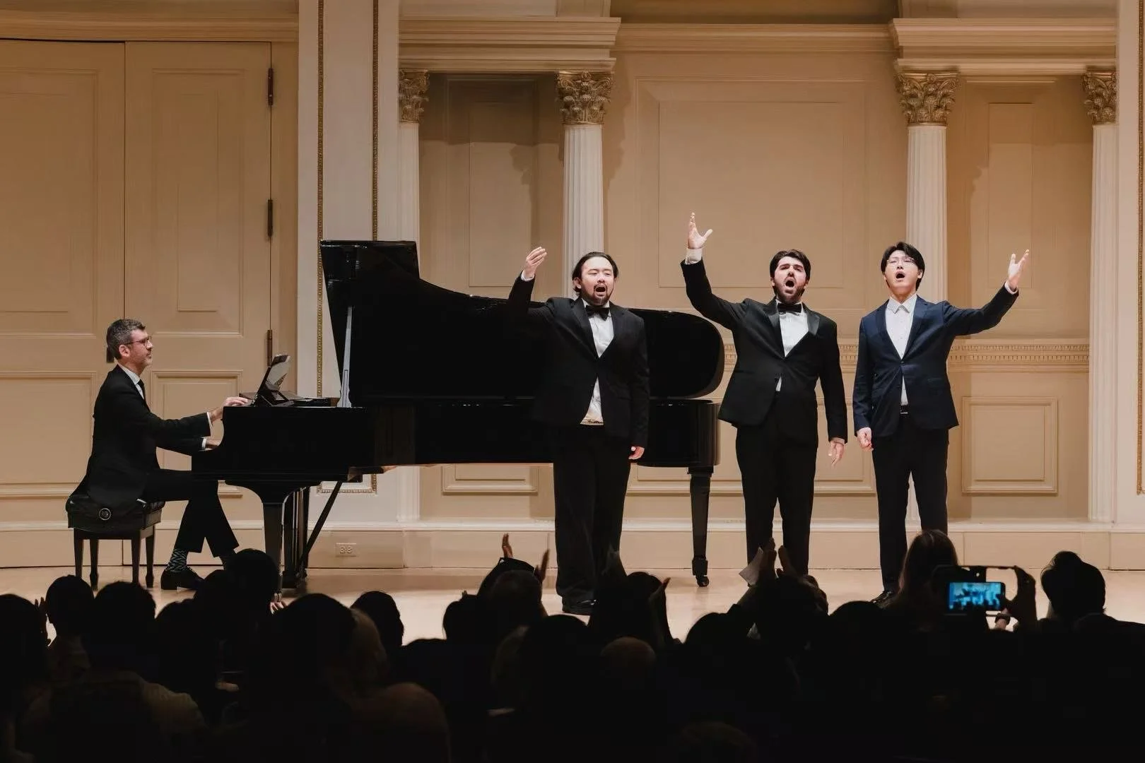 A pianist playing a grand piano while three men in tuxedos sing on stage during a concert. Audience members are visible in the foreground, some taking photos.