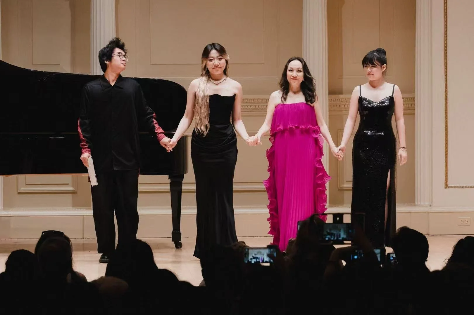 Four performers on stage holding hands, wearing formal attire, with a grand piano in the background and audience members taking photos.