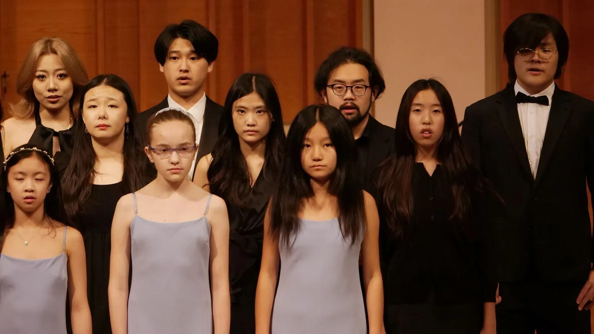 A group of people, including young girls and adults, dressed in formal attire, standing in front of wooden paneled walls, possibly at a concert or event.