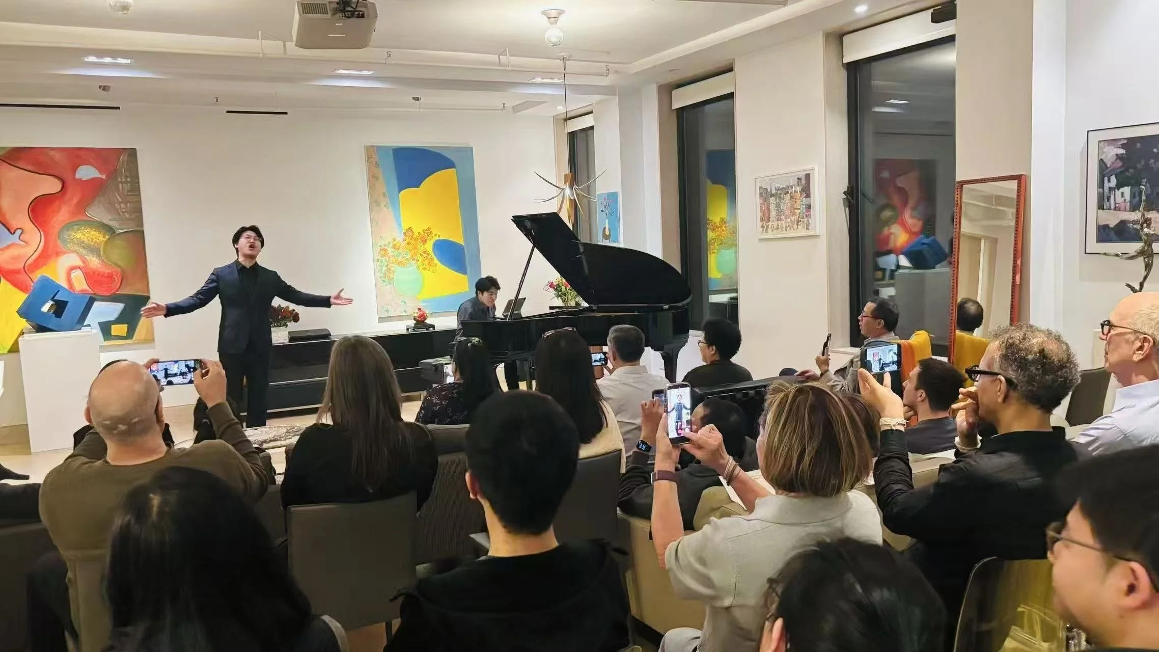 A man is singing or performing in front of an audience in an art gallery or exhibition space. There is a grand piano and various colorful artwork on the walls. Audience members are seated and capturing the moment with their phones.