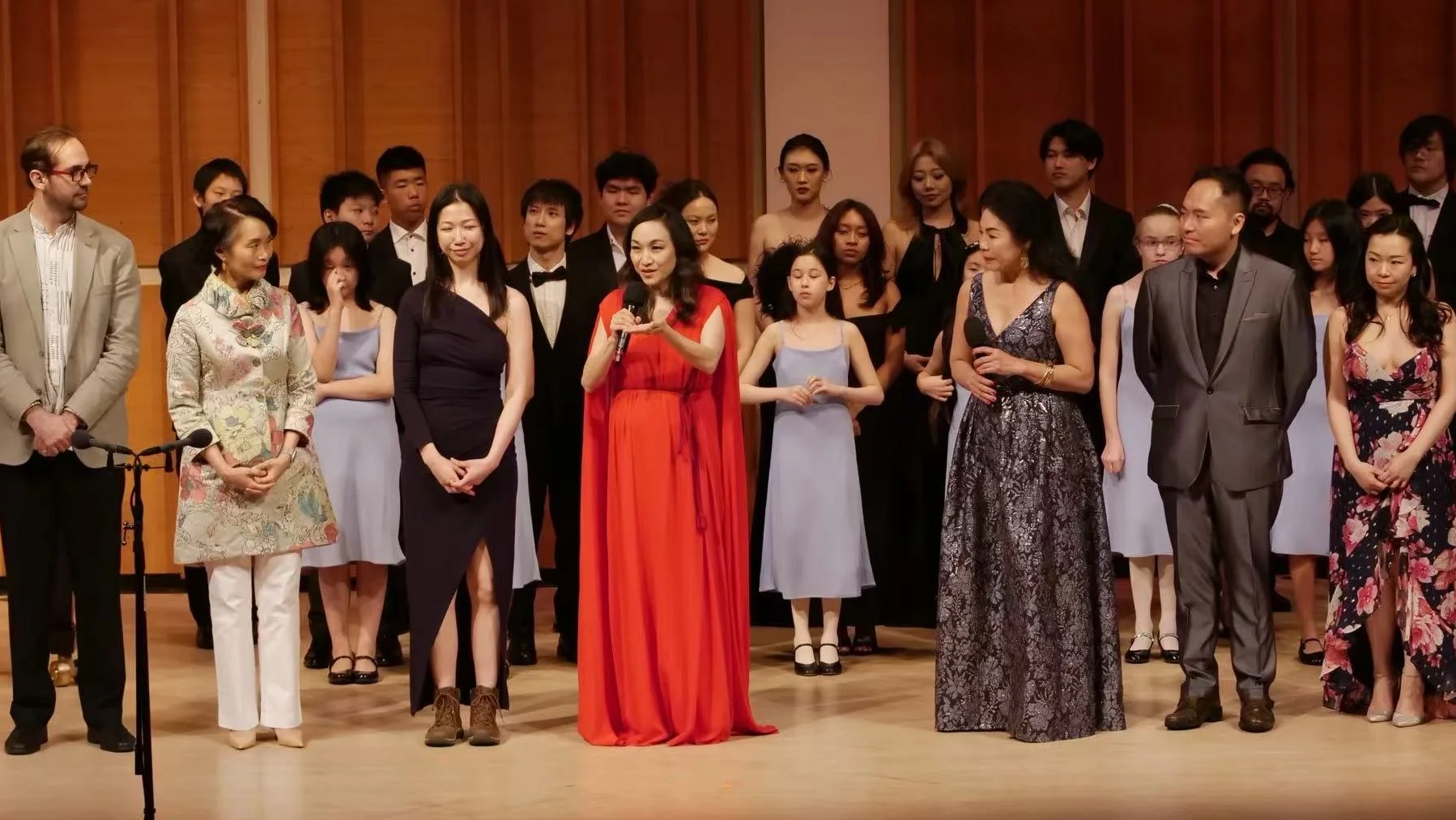 Group of people on stage, including women and men dressed in formal and semi-formal attire, some holding microphones, standing against a wood-paneled backdrop, at a formal event or performance.