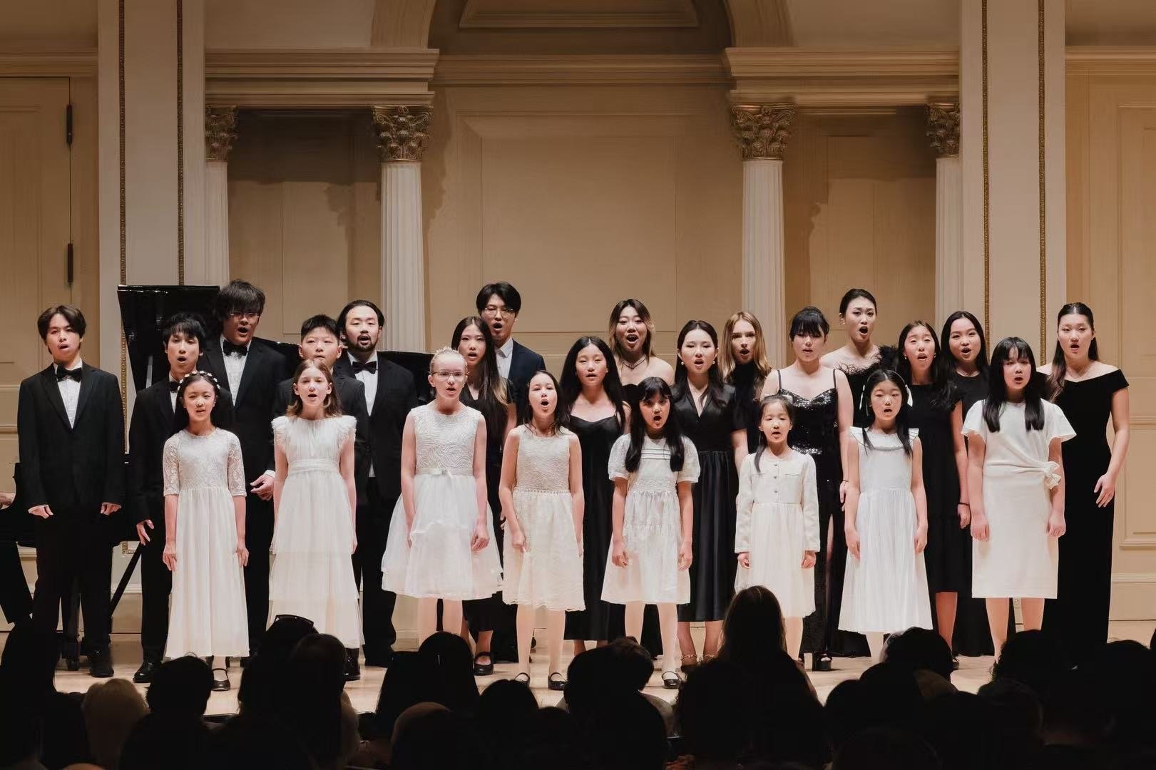 Children and young adults singing in a concert hall, dressed in formal attire, with some wearing black and others in white dresses and suits.