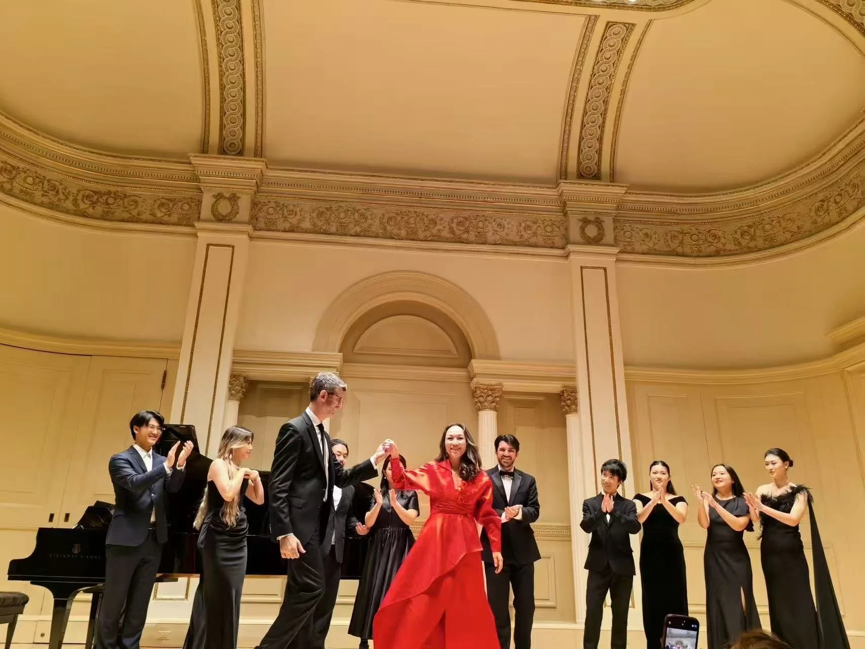 A group of people dressed in formal attire standing on a stage in a grand, ornate room with high ceilings and gold decorations. One woman in a red gown is at the center, holding hands with a man in a black suit and tie. The audience members are clapping and smiling.
