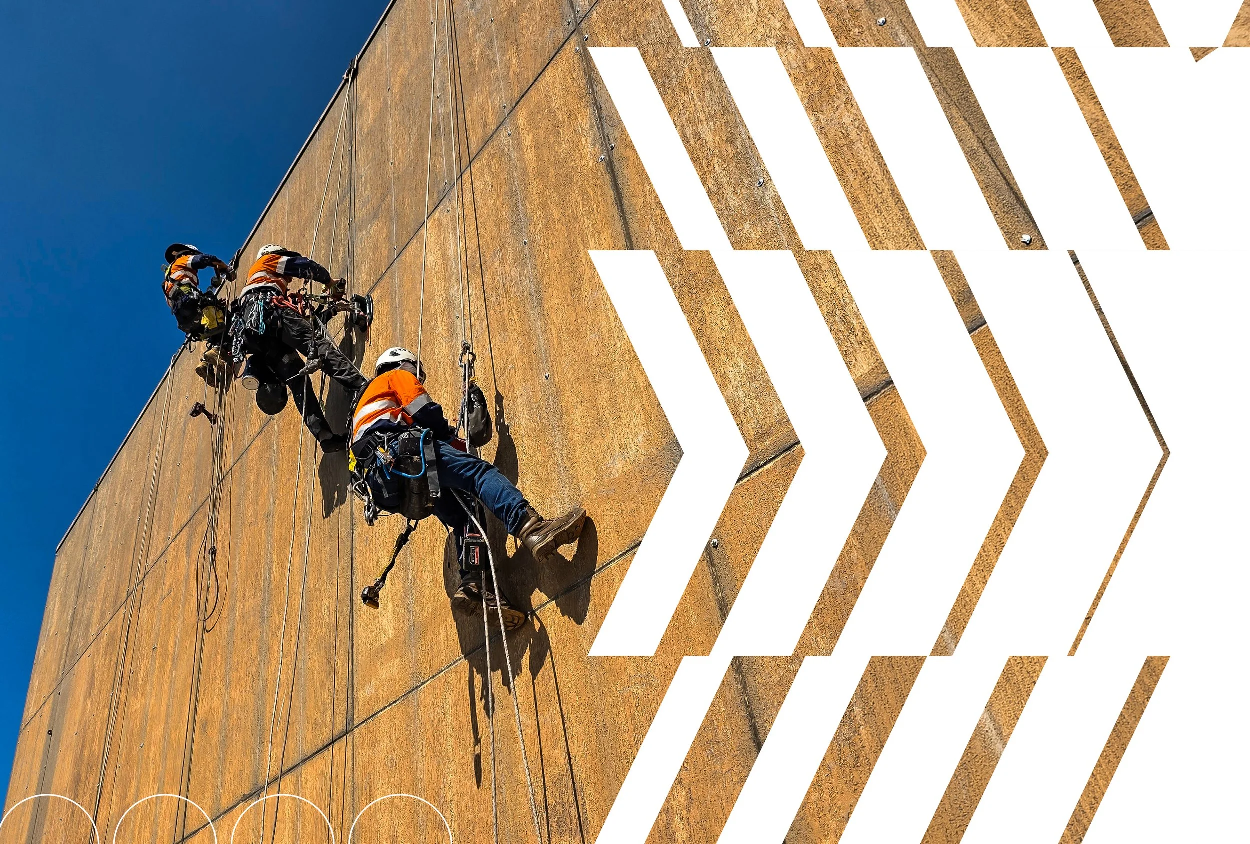Two workers climbing a large vertical metal wall during construction or maintenance, wearing safety helmets and harnesses, with blue sky in the background.