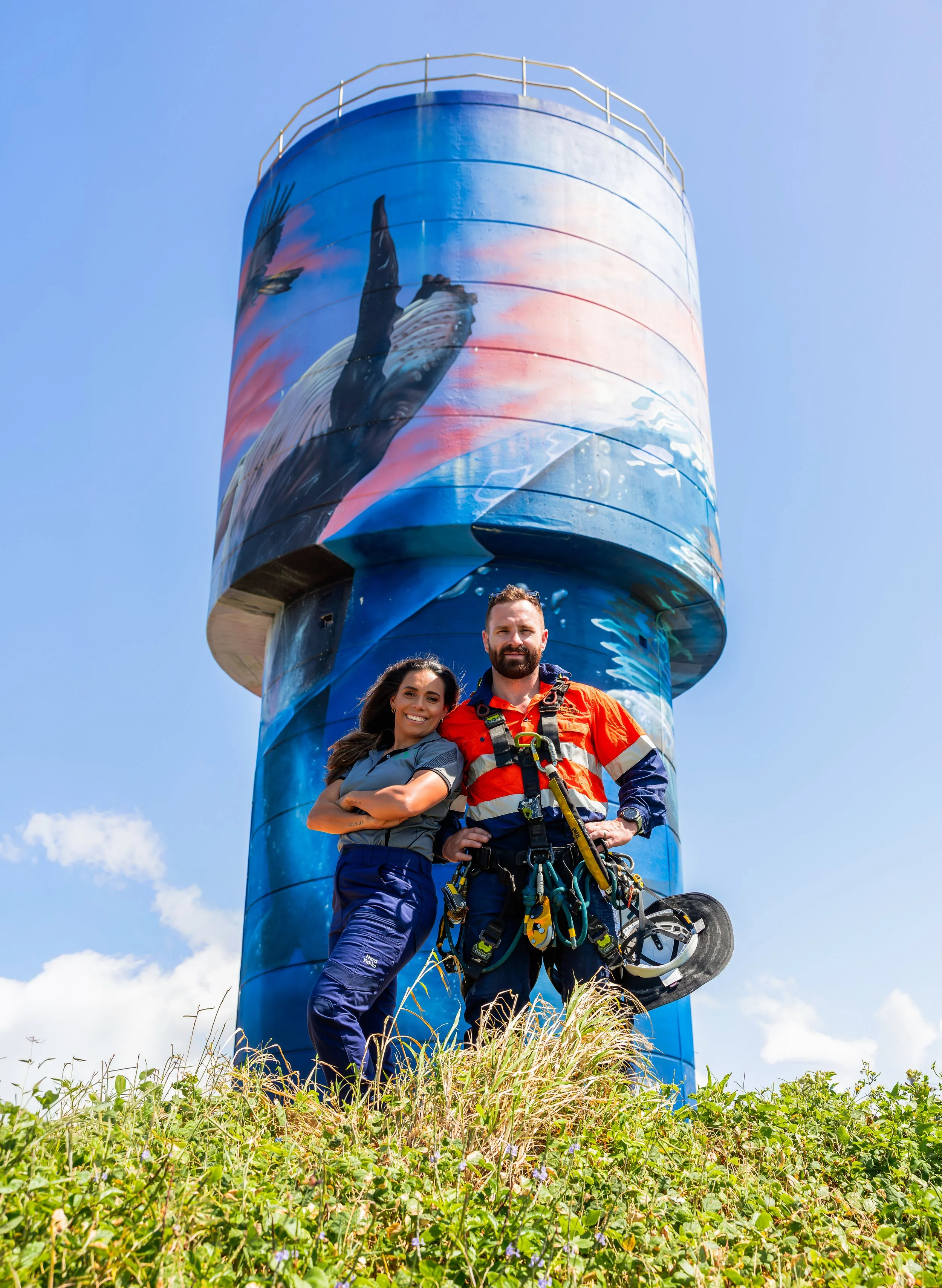 Two people are standing in front of a tall, cylindrical tower with a colorful mural of an ocean scene, including a whale and a bird. The man is dressed in outdoor gear, holding a helmet, and the woman is smiling with her arms crossed. The sky is clear with a few clouds and the ground is covered with grass and small plants.