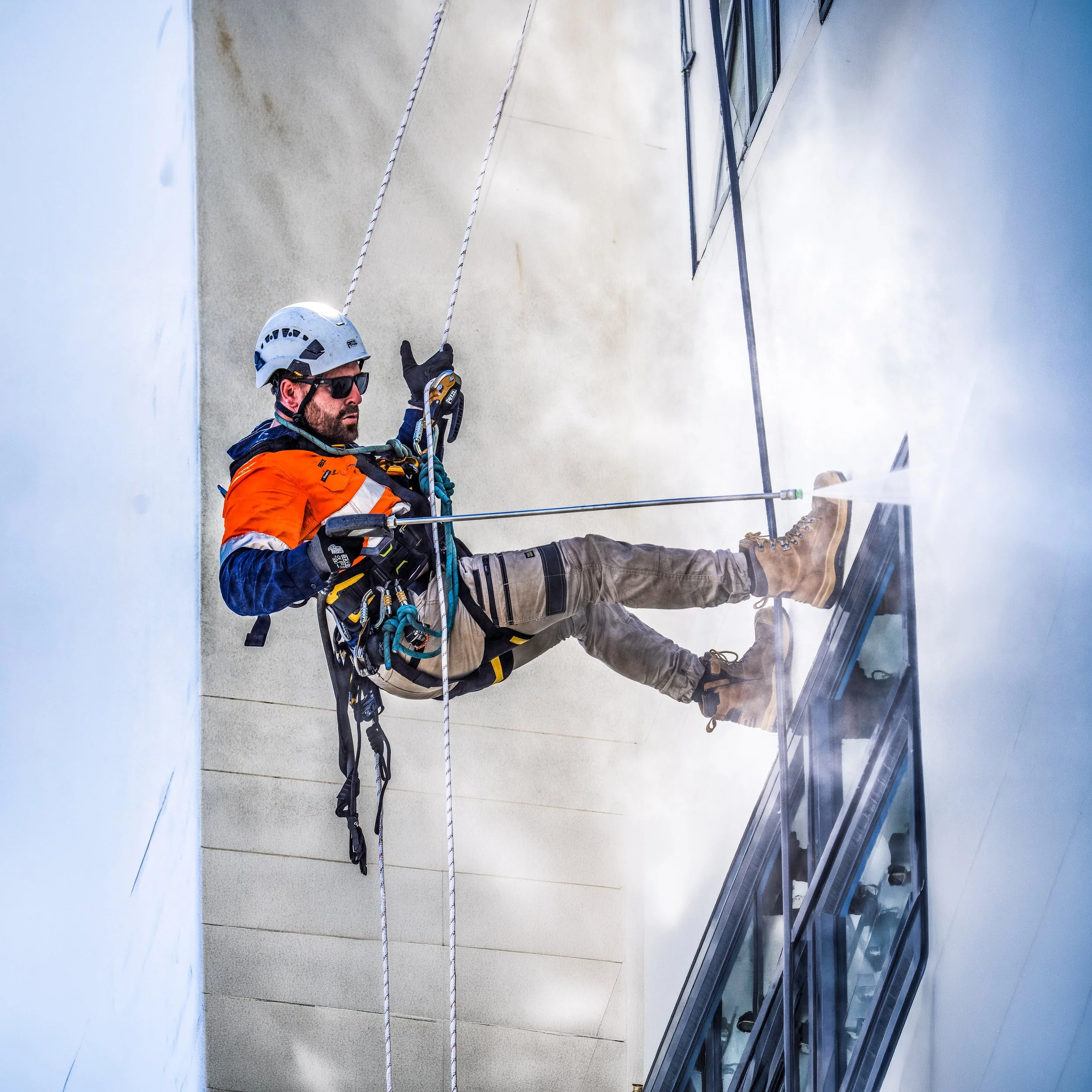 A man wearing a safety helmet, sunglasses, and work gloves is using climbing equipment to perform maintenance on the exterior of a building, with a clouded sky in the background.