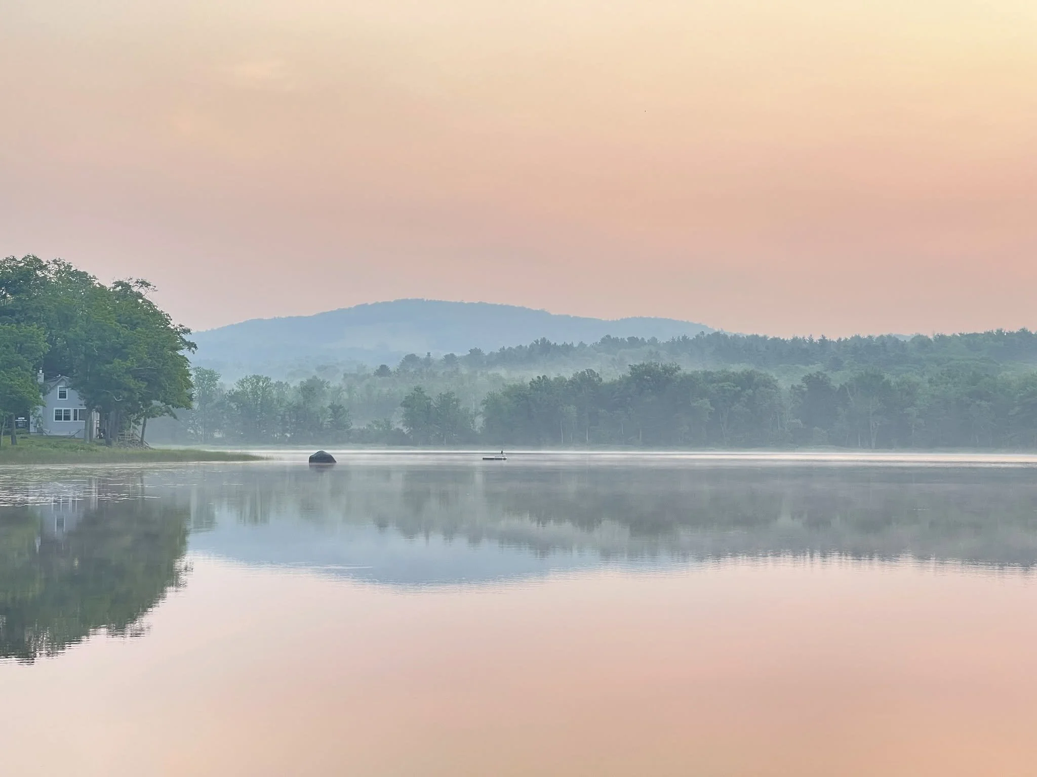 A calm river with a small boat floating on it, surrounded by green trees and distant hills under a softly colored sky.