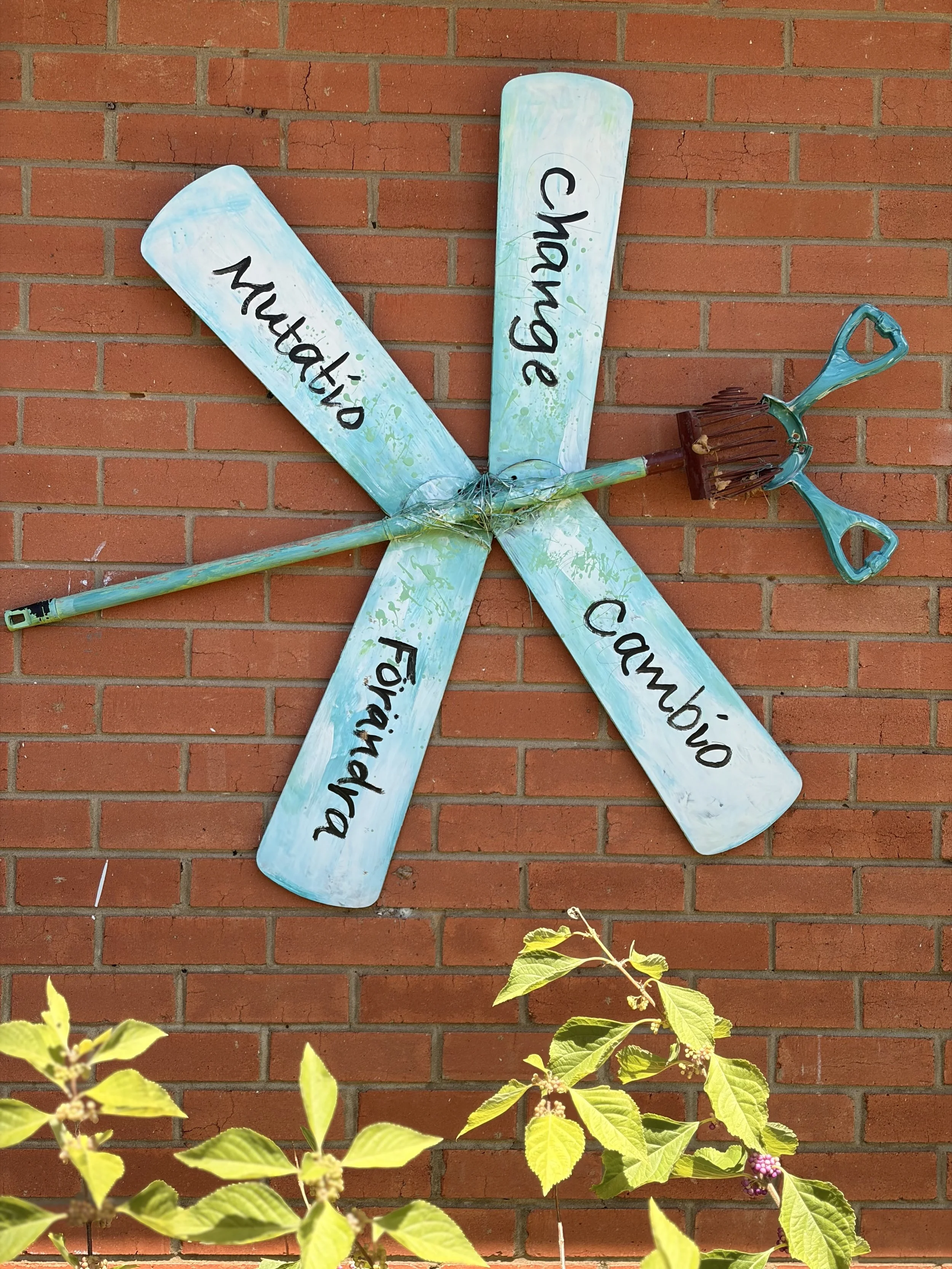 A decorative wall hanging of a greenish blue-painted dragonfly with four wings, each bearing the words: "change" in four different languages. english, spanish, italian, and swedish.  made out of ceiling fan parts and the main body is a pecan picker u