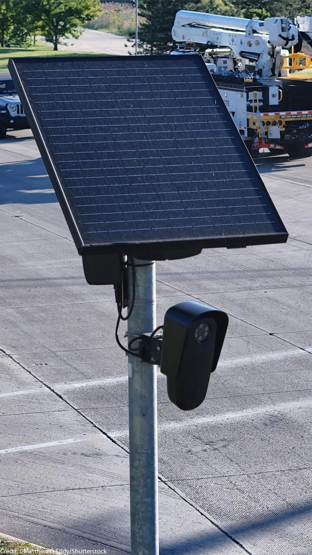 A solar panel mounted on a pole in a parking lot, with a security camera attached below the panel and a utility truck with a lift in the background.