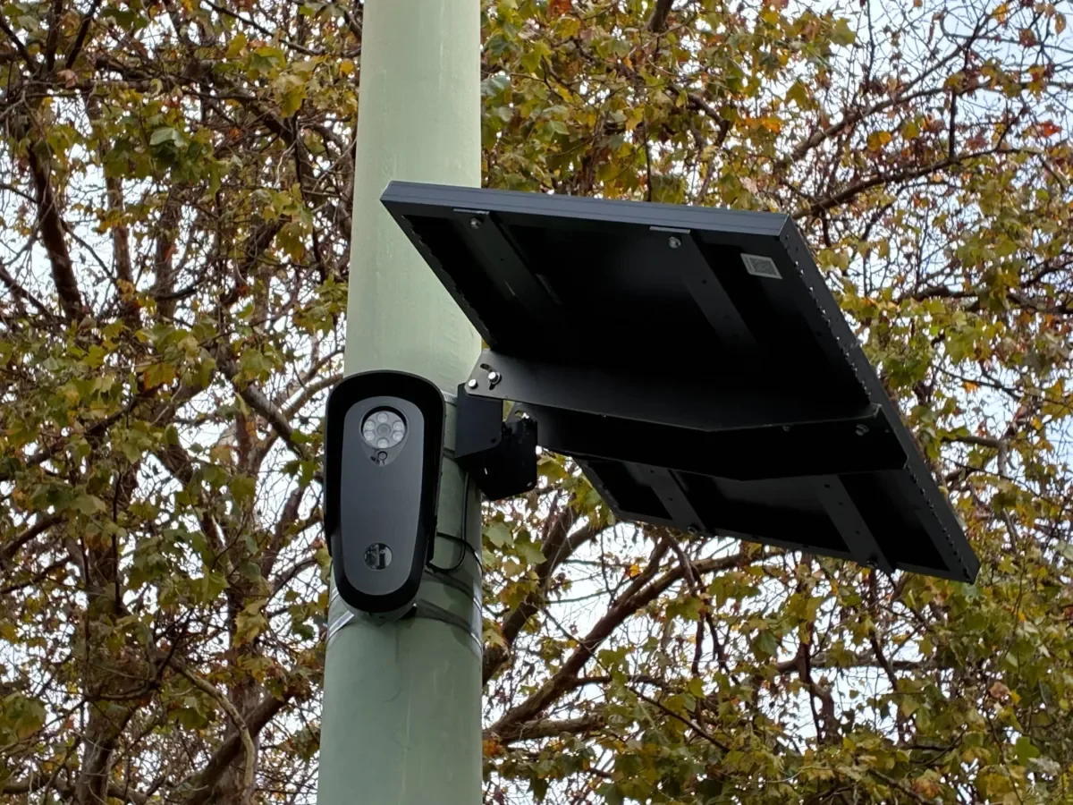 A surveillance camera and solar panel mounted on a green utility pole with trees in the background.