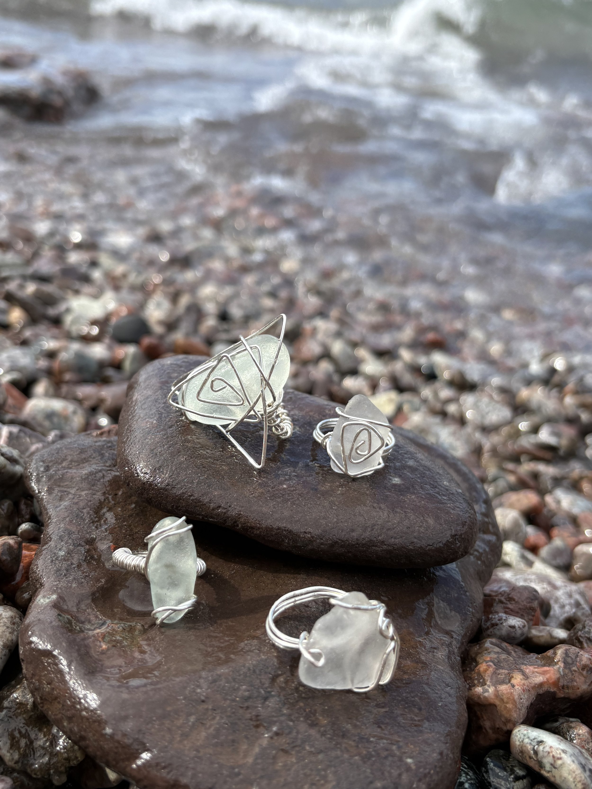 Jewelry with sea glass and wire designs on rocks at a shoreline with waves and pebbles.