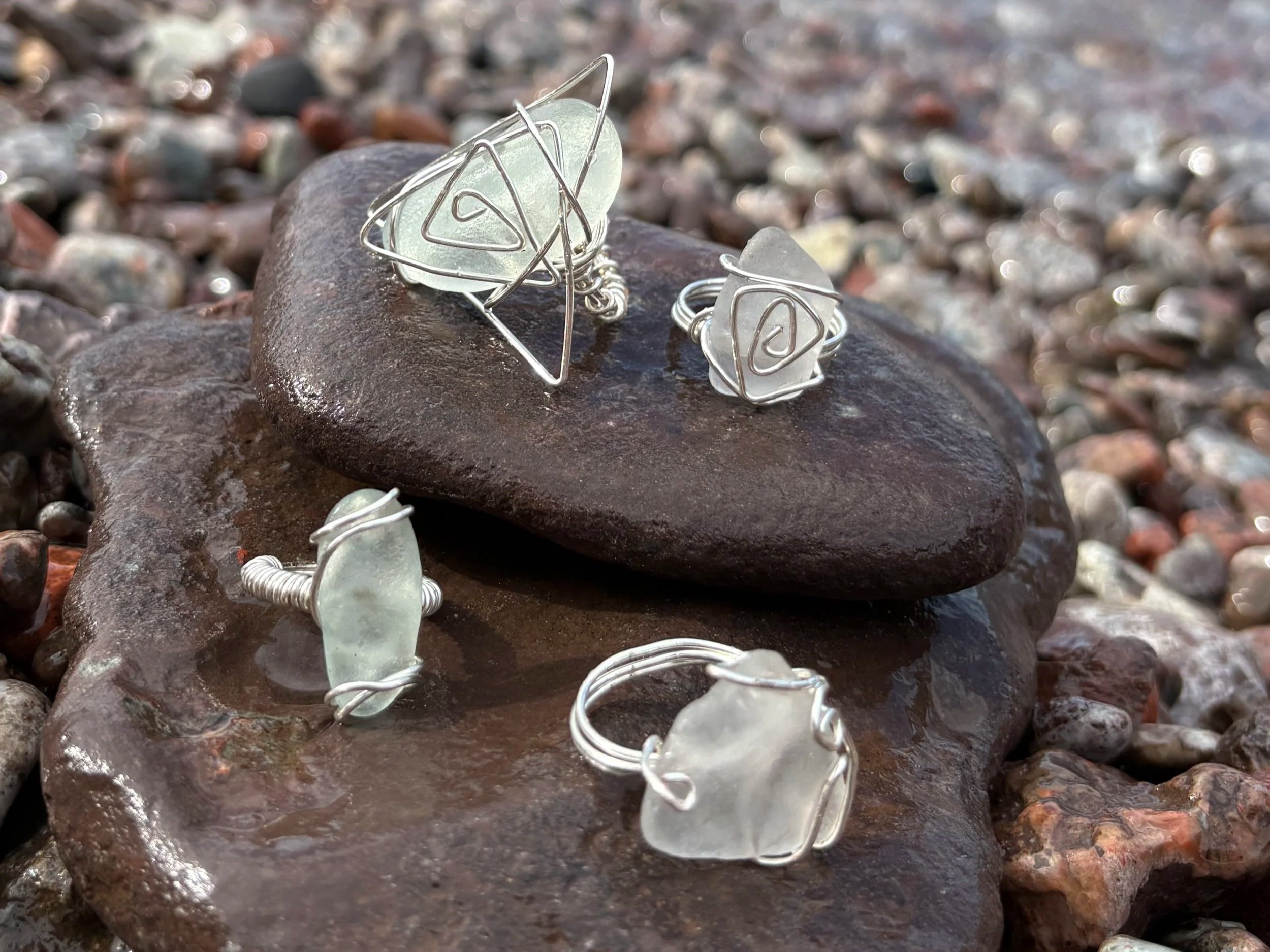 Silver wire jewelry with sea glass pieces placed on wet rocks by the shoreline.