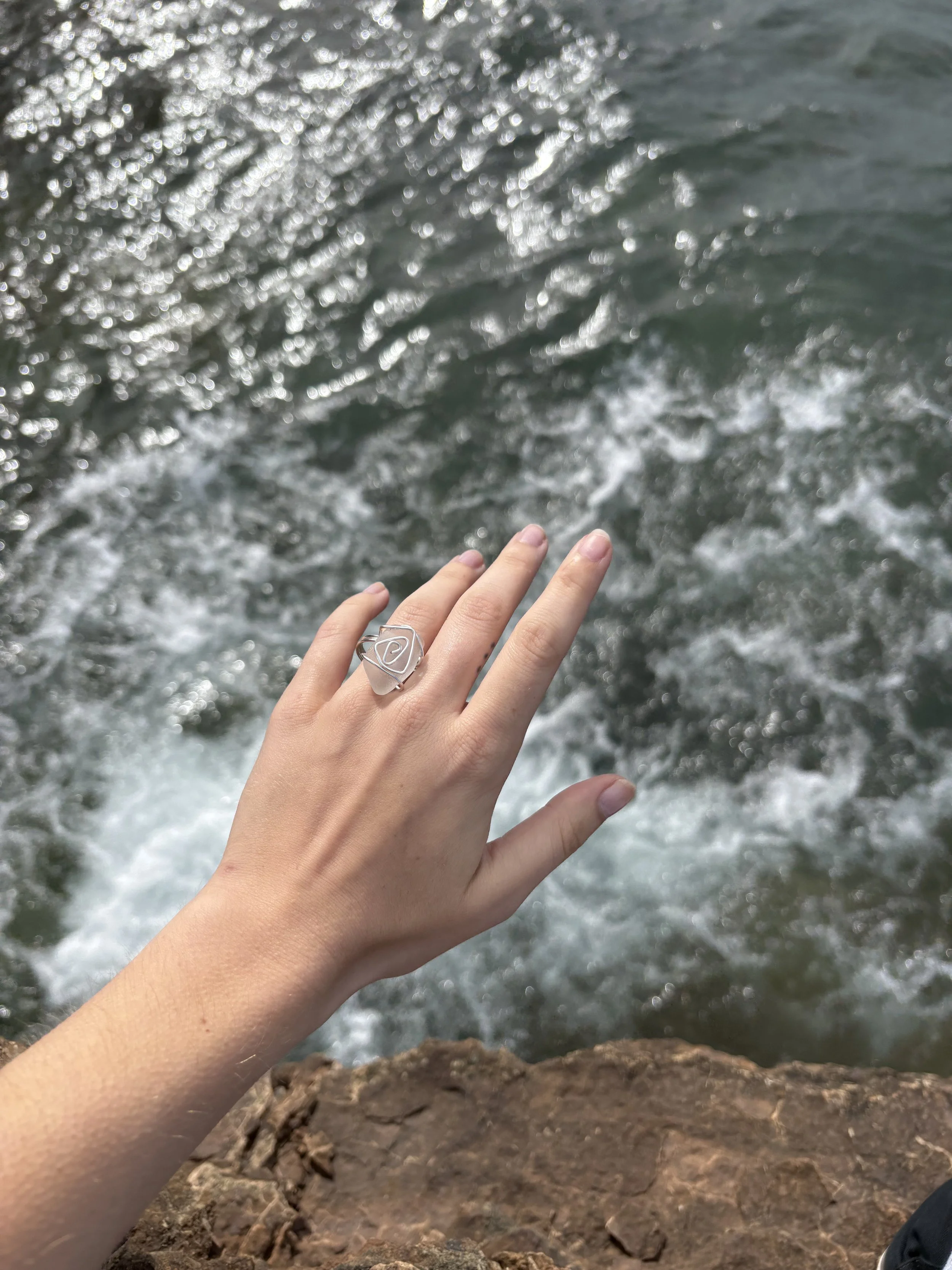 A person's hand with a silver ring near a body of water with waves and foam, with a rocky shore in the foreground.