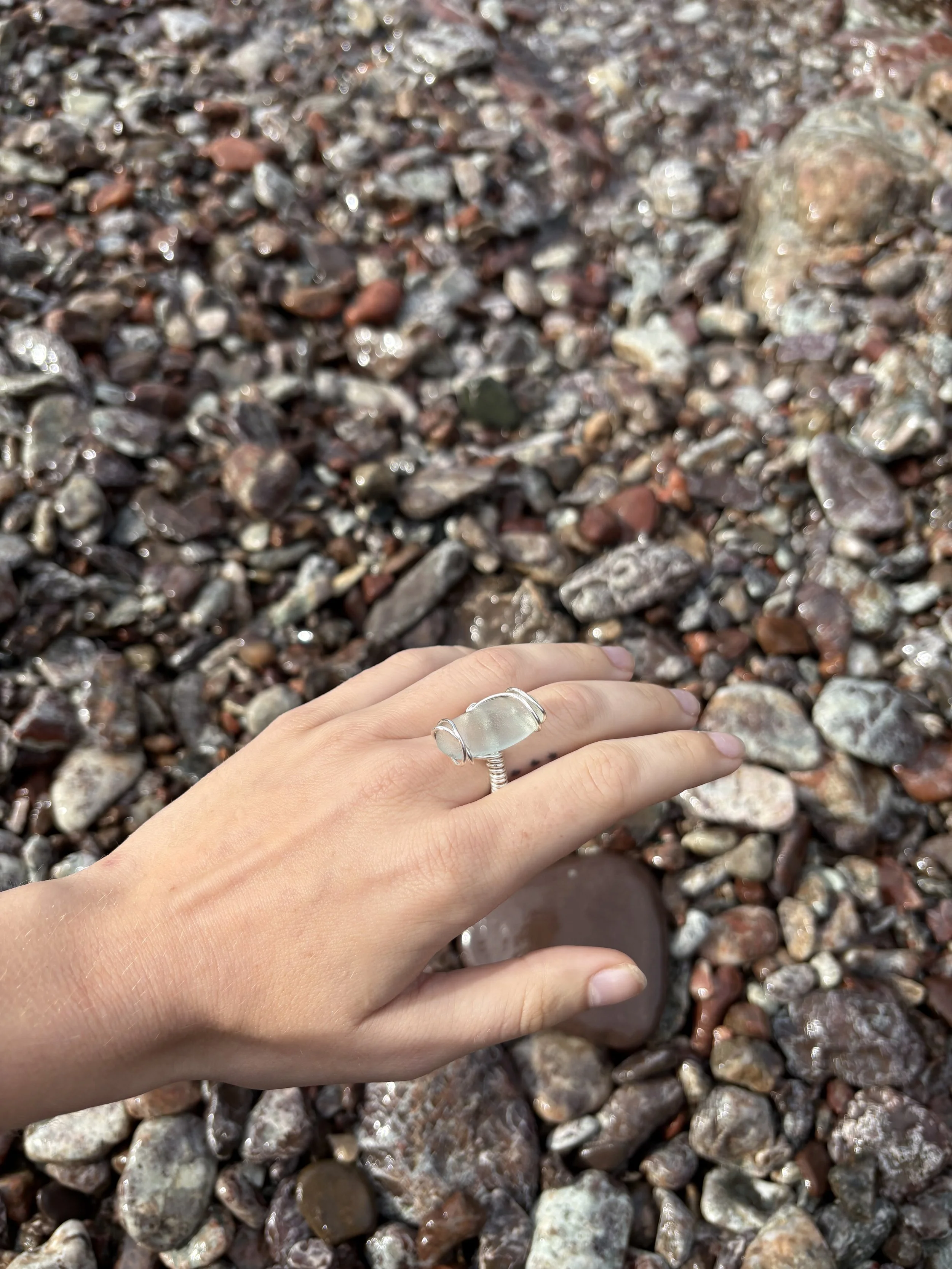 Close-up of a hand with a silver ring touching wet, multicolored small rocks and pebbles on a beach or riverbank.
