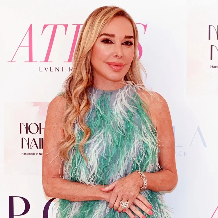 A woman with long wavy blond hair and light skin, wearing a sleeveless dress with feather details in shades of green and white, standing in front of a white backdrop with pink and black text.