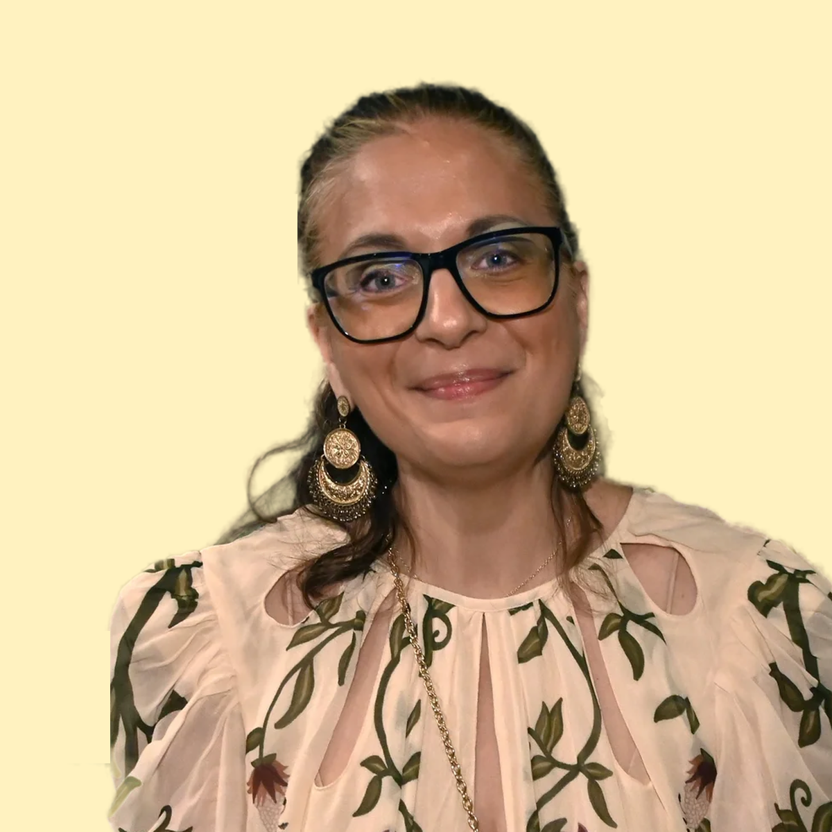 A woman with glasses, long dark hair, and earrings, smiling at the camera, wearing a light-colored floral blouse, with a plain pale yellow background.