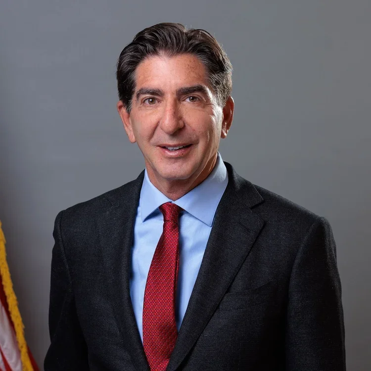 A man with dark hair, wearing a gray suit, light blue shirt, and red tie, standing in front of a gray background with part of an American flag visible on the left.
