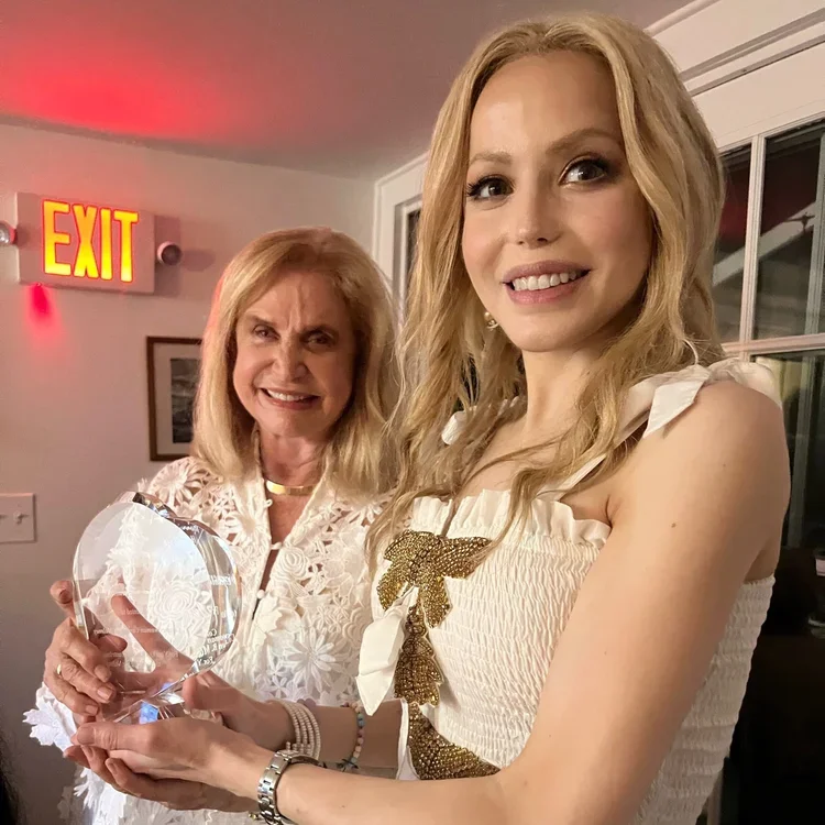 Two women standing indoors, smiling at the camera. The older woman on the left has blonde hair and is holding a clear glass award. The younger woman on the right, with long blonde hair, is wearing a white dress with gold embellishments and is assisting with the award.