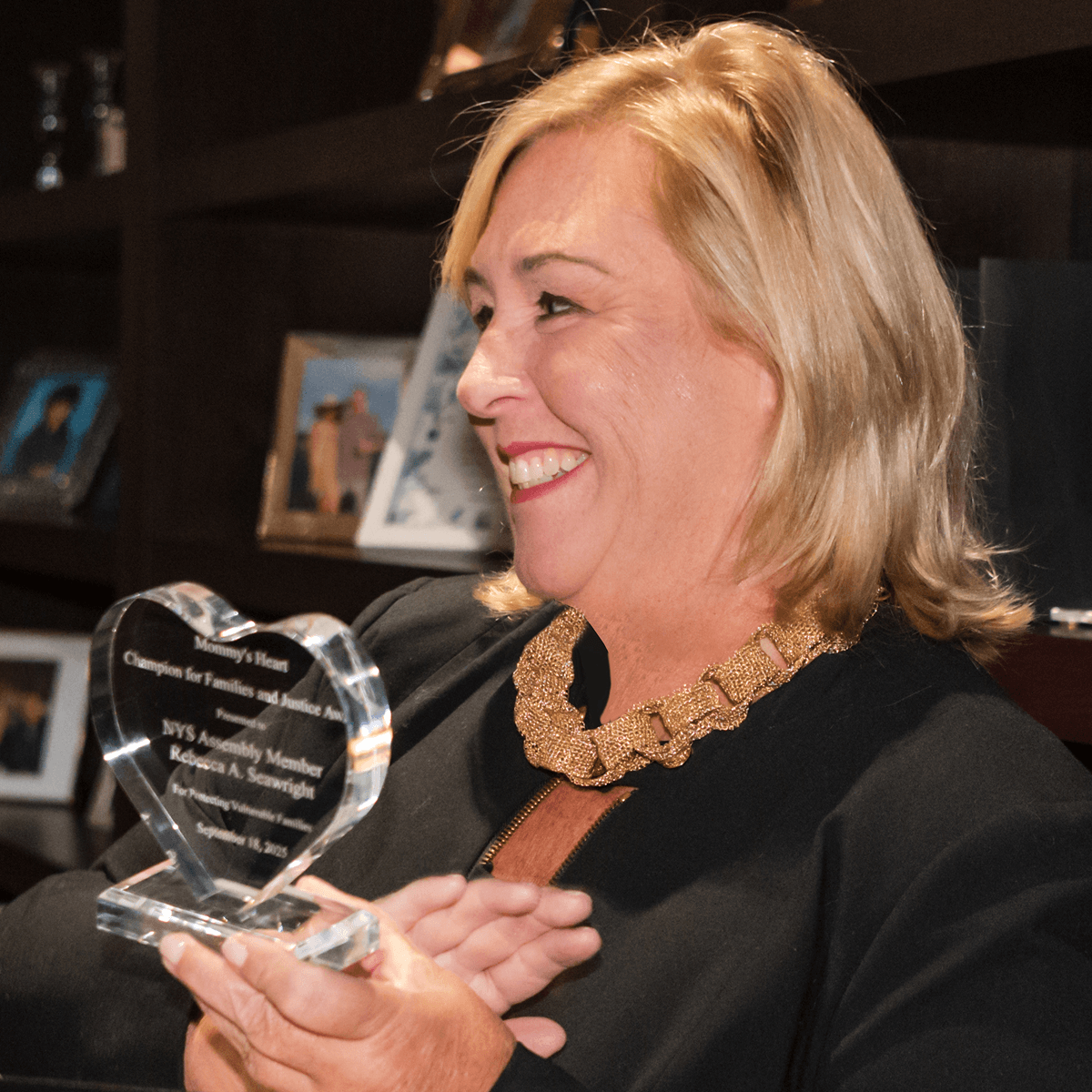 A woman with blonde hair smiling and holding a clear heart-shaped glass award. She is wearing a black outfit with a gold chain necklace, standing in front of a dark shelf with framed photos.