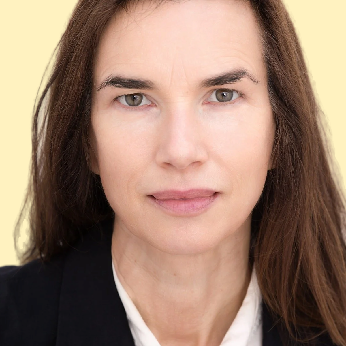 Close-up portrait of a woman with long brown hair, wearing a black blazer and white shirt, against a light yellow background.
