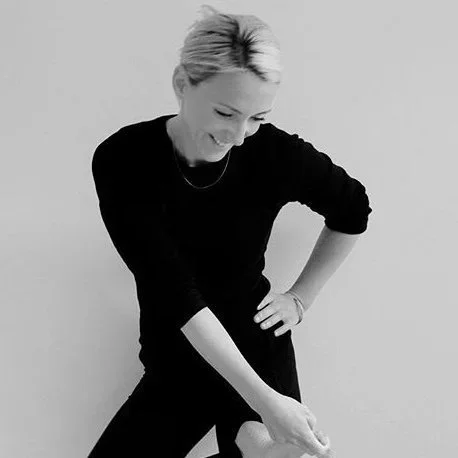 Black and white photo of a woman with short blonde hair smiling and looking down, wearing a black long-sleeve top and pants, with one hand on her hip and the other resting on her thigh.