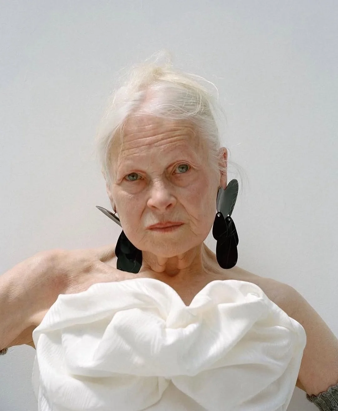 An elderly woman with white hair and large black and gray earrings, wearing a white off-shoulder top, looking directly at the camera with a neutral expression.
